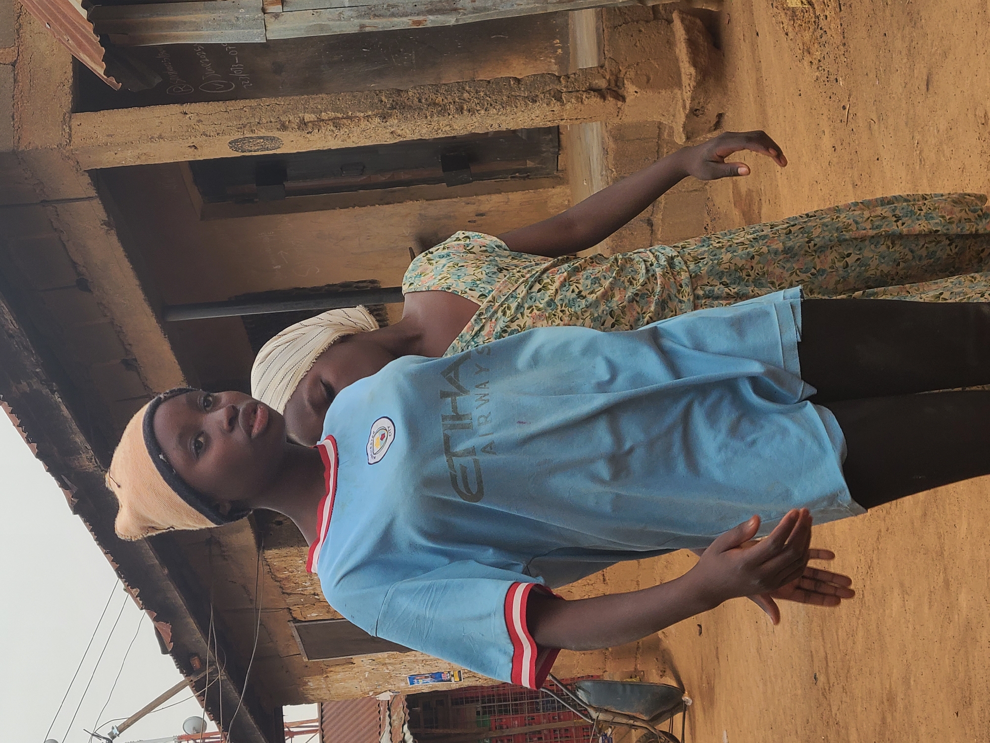Two young people stand outside a building. The person in the foreground wears a light blue Manchester City football jersey with a red and white striped collar. A light brown cap covers most of their hair. The second person, slightly behind, wears a floral print dress and a white striped head covering. The background shows a building with a corrugated metal roof and a dirt ground. A bicycle and stacks of red crates are visible near the ground. The location is Jos, Nigeria.