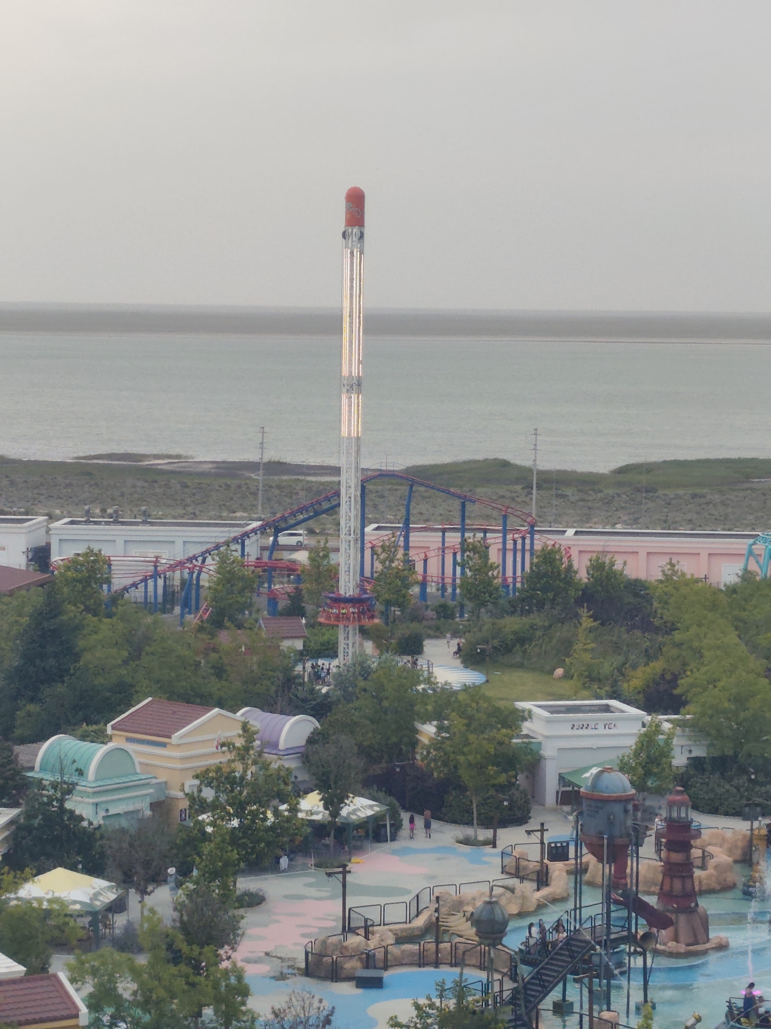An aerial view captures an amusement park situated by a body of water, likely the Caspian Sea, in Aktau, Kazakhstan. The park features a tall, slender tower ride with lights, a roller coaster with red and blue tracks, and various buildings with colorful roofs. Green trees are scattered throughout the park, and a large body of water is visible in the background, separated from the park by a strip of land. Several people are present, some walking on paths, others near attractions. The overall scene depicts a recreational area in an urban or coastal setting.