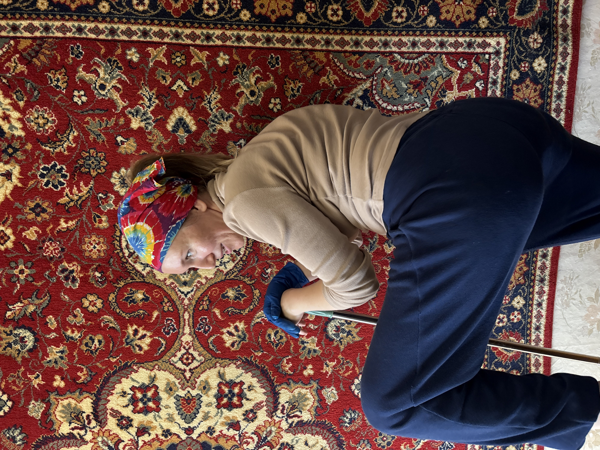 A person lies on a red Persian rug, possibly cleaning or performing some task. They are wearing a beige sweatshirt, dark blue sweatpants, and blue gloves. A colorful bandana is tied around their head. The person is holding a cleaning tool, possibly a mop or broom, with a metal handle. The rug has a traditional pattern with red, blue, yellow, and white colors. The scene is indoors, with part of a light-colored wall visible to the right. The lighting suggests it could be daytime. The person's expression is neutral. There is no visible text in the image.