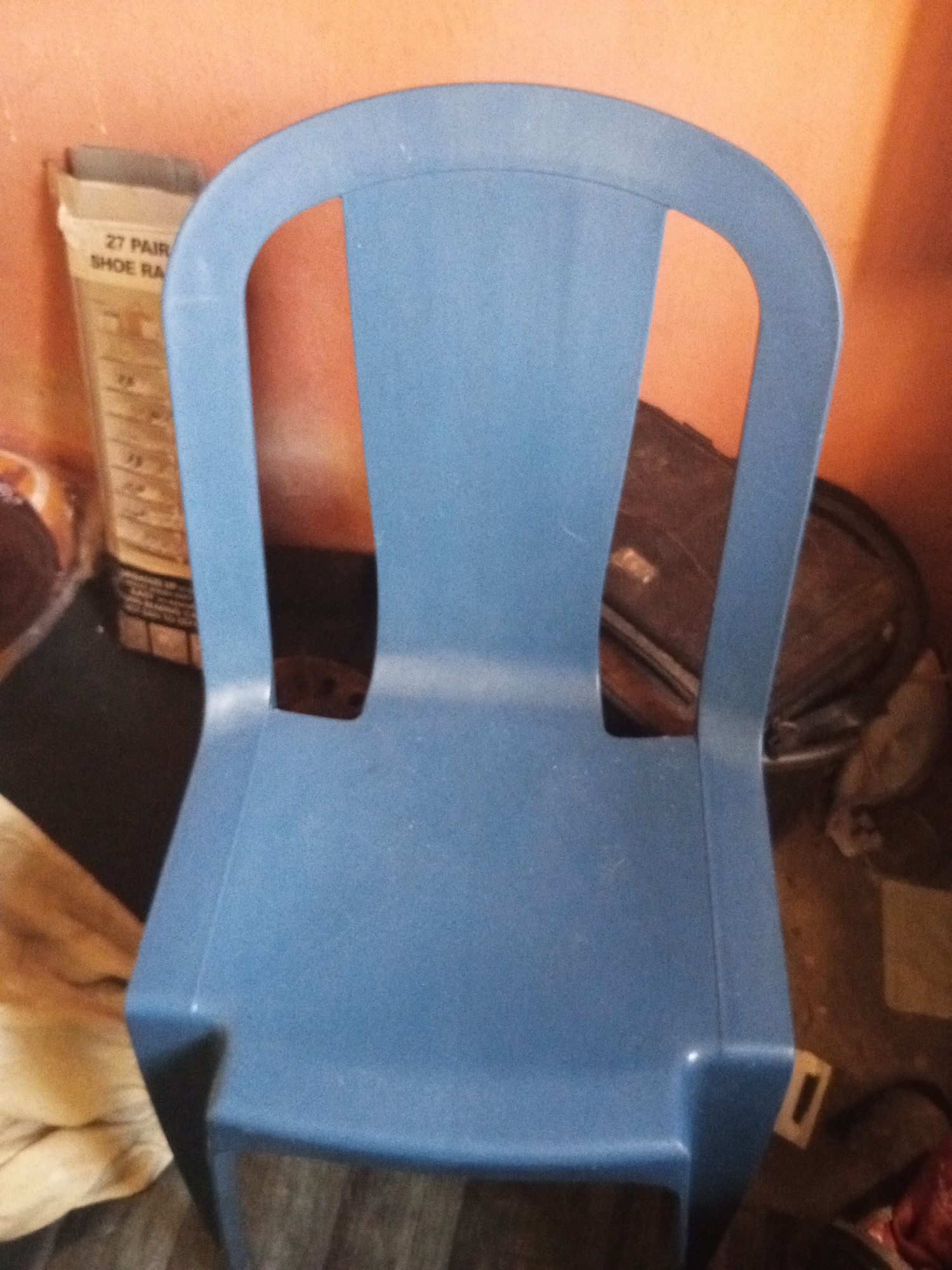A blue plastic chair is positioned in the foreground, facing forward. Behind the chair, to the left, a cardboard box with text that appears to read "27 PAIR SHOE RA" is visible. The background consists of an orange-colored wall and a dark brown object, possibly a drum or barrel, with a circular lid. To the left of the chair, a beige fabric object is draped. The flooring appears to be a dark material. The scene is set indoors.