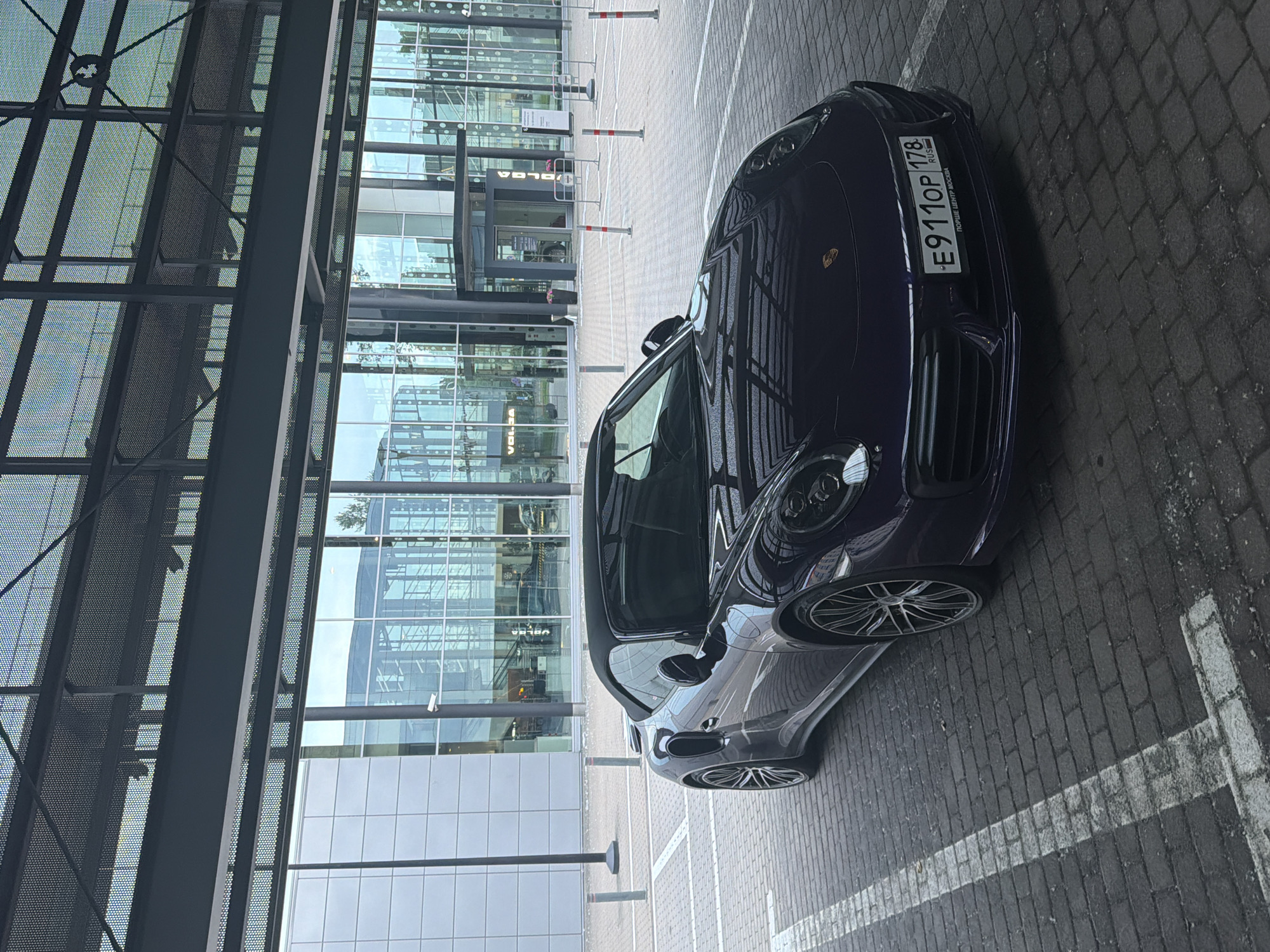 A dark purple Porsche 911 convertible is parked on a brick-paved surface under an overhang. The car is facing forward, with its front bumper visible and a license plate that reads "E911OP 178 RUS". The background features a modern glass-fronted building with a dark metal structural framework. The building has large windows reflecting the sky and surrounding environment. Several bollards are visible in the foreground and midground. The lighting suggests an overcast day.