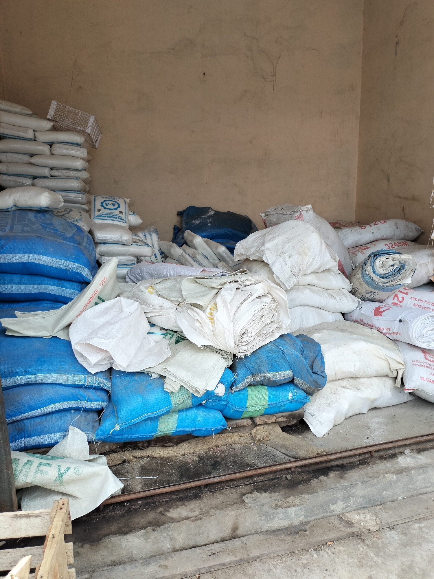 In a dimly lit storage area in Kotapantonlabu, Indonesia, a large quantity of bagged goods are piled high. The bags, made of woven plastic and burlap, are stacked precariously against a textured, light brown wall. The floor is a rough, uneven concrete surface with some debris and a metal pipe running across it.
The majority of the bags are white or light-colored, with some featuring blue or green stripes. Several bags are labeled with text, including one with the "A-A" logo and other illegible Indonesian script. Some bags appear to be tightly packed and sealed, while others are loosely filled or partially open, with their contents potentially exposed.
In the foreground, a portion of a wooden crate is visible. The overall impression is one of a storage space filled with raw materials or products, possibly for agricultural or industrial use. The lighting is dim, suggesting an indoor or covered area, and the environment appears somewhat unkempt. There are no people visible in the image.
