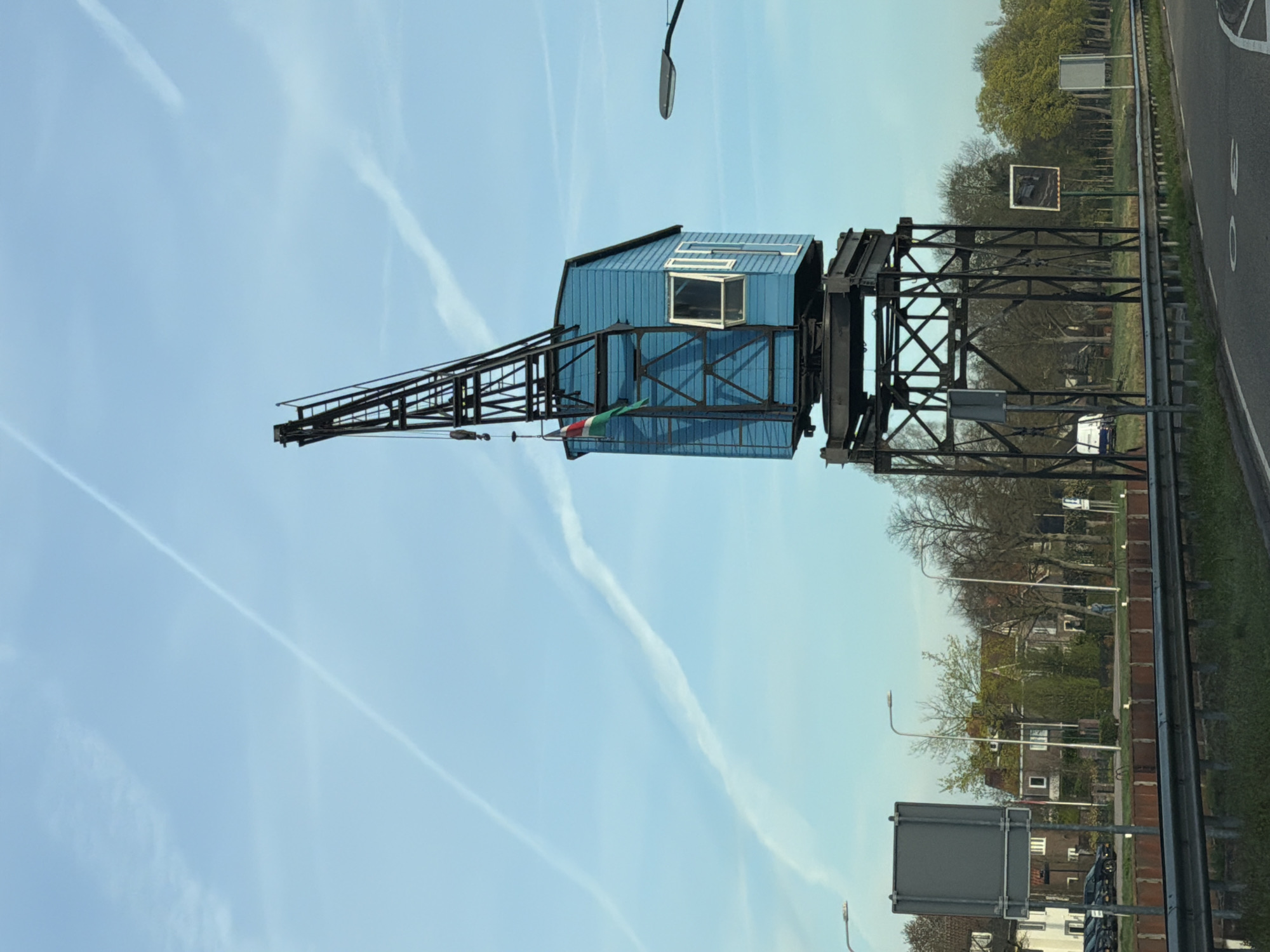 A horizontal photograph captures a blue crane against a light blue sky streaked with contrails. The crane's cab is painted blue, with a white-framed window slightly ajar. A flag with red, white, and green stripes is visible near the crane's arm. The crane is constructed of black metal and stands on a sturdy base. A road runs alongside the crane, with a metal guardrail visible. Trees and houses can be seen in the background. Streetlights stand on the other side of the road. The scene is in Beek en Donk, Netherlands.
