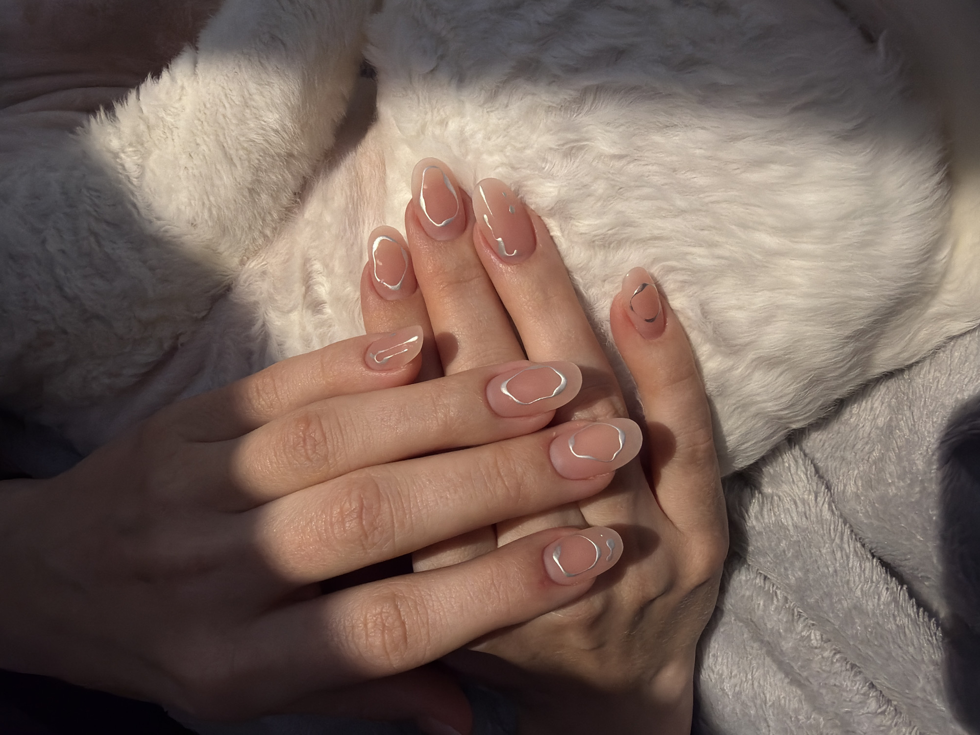 A pair of female hands with manicured nails is resting on soft blankets. The nails are oval-shaped with a nude base and a silver abstract design. The background consists of a white fluffy blanket in the upper portion and a grey plush blanket in the lower portion. The lighting is a mix of sunlight and shadow, suggesting an indoor setting. There is no visible indication of the location being Ramat Gan, Israel, or any other specific place. The image focuses primarily on the hands and the nail art.