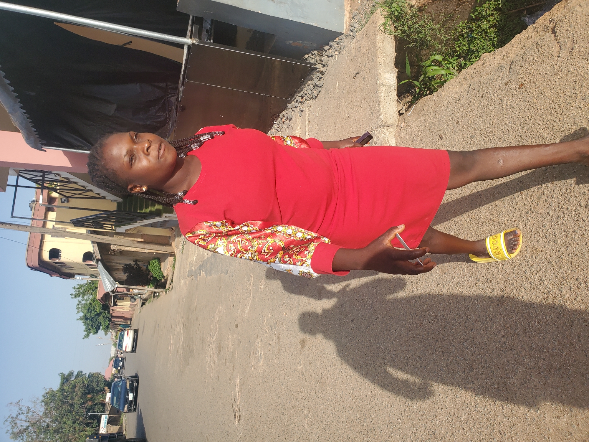A woman stands on a street in Jos, Nigeria. She wears a red dress with patterned sleeves and yellow sandals. She holds a cell phone in one hand and a small, possibly metallic, object in the other. She has braided hair. The street behind her shows buildings, parked cars, and vegetation. The background suggests an urban area with residential and commercial structures. The lighting indicates it is likely daytime with direct sunlight.