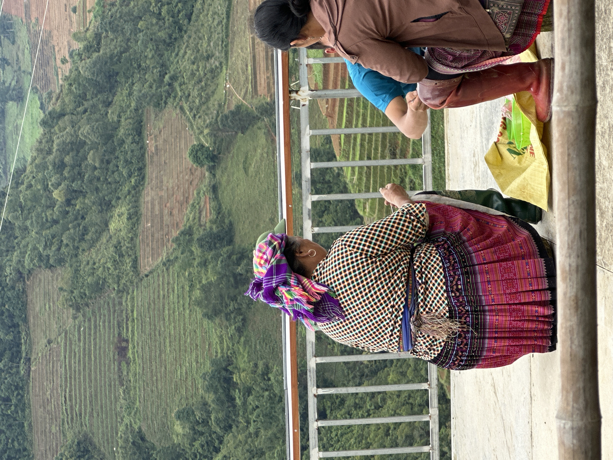 Three individuals are seated on a concrete surface, observed from the back, overlooking a terraced mountainous landscape in Mù Cang Chải, Vietnam. A silver metal railing with a wooden top bar spans the mid-ground. The person on the left wears a vibrant, multi-colored plaid headscarf (purple, pink, green, yellow), a black, white, and orange plaid shirt, a blue waistband with tassels, and a long pleated skirt featuring intricate pink, red, and black patterns. A gold hoop earring is visible on their right ear, and a dark green rubber boot is partially seen. Their hands are positioned in front, holding a small, light-colored object. A second person, a male in a bright blue t-shirt, is partially visible in the center. The third person, on the right, has dark hair tied back and wears a brown jacket and a patterned skirt with tall reddish-brown rubber boots. Between the central and right figures, a yellow fabric bag rests on the ground, containing two light green plastic bowls. The background displays extensive mountainous terrain covered in lush green foliage and numerous rice terraces, some vibrant green with growth and others showing exposed reddish-brown earth, under an overcast sky. Narrow dirt paths are visible winding through the distant hillsides.