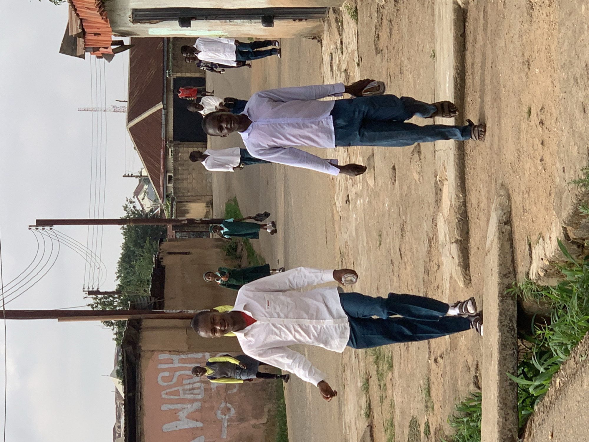 The image shows a street scene in Jos, Nigeria. Two young men in white shirts and blue pants are standing on the side of a paved street. Other individuals, possibly students in uniforms, are walking in the background. Buildings line the street. Power lines are visible overhead. The setting appears to be a residential or commercial area.