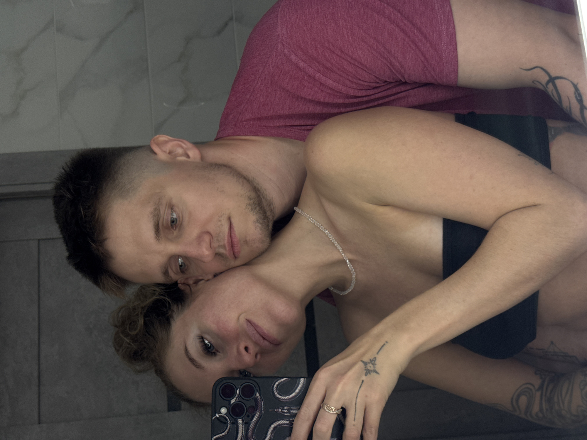 A man and a woman are taking a selfie in a bathroom. The woman is in the foreground, holding a smartphone with a snake-patterned case and a ring on her finger. She has her hair pulled up and is wearing a black strapless top and a beaded necklace. The man is behind her, with his arm around her. He is wearing a pink t-shirt. The background appears to be a tiled wall. The image does not provide any information about the city of Chaikovsky, Russia.