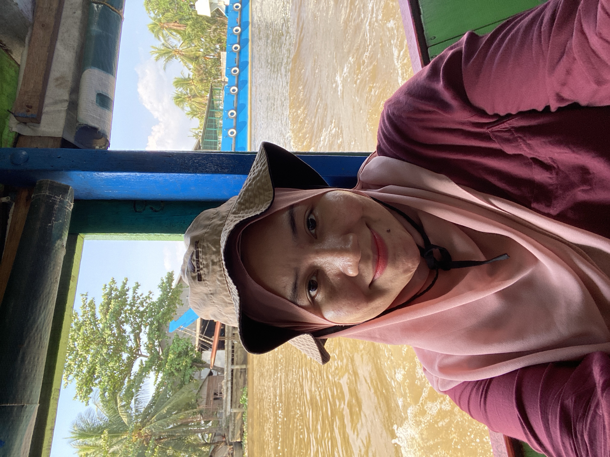 A medium shot photograph captures a woman wearing a pink hijab and a maroon long-sleeved shirt, positioned within a boat. She is also wearing a light brown bucket hat. The woman is smiling directly at the camera. The boat appears to be on a river, with murky brown water visible. In the background, palm trees and vegetation line the riverbank, and several buildings are visible. A bright blue boat with white circular markings is also present on the river. The scene suggests a location in or around Banjarmasin, Indonesia, a city known for its extensive river network.