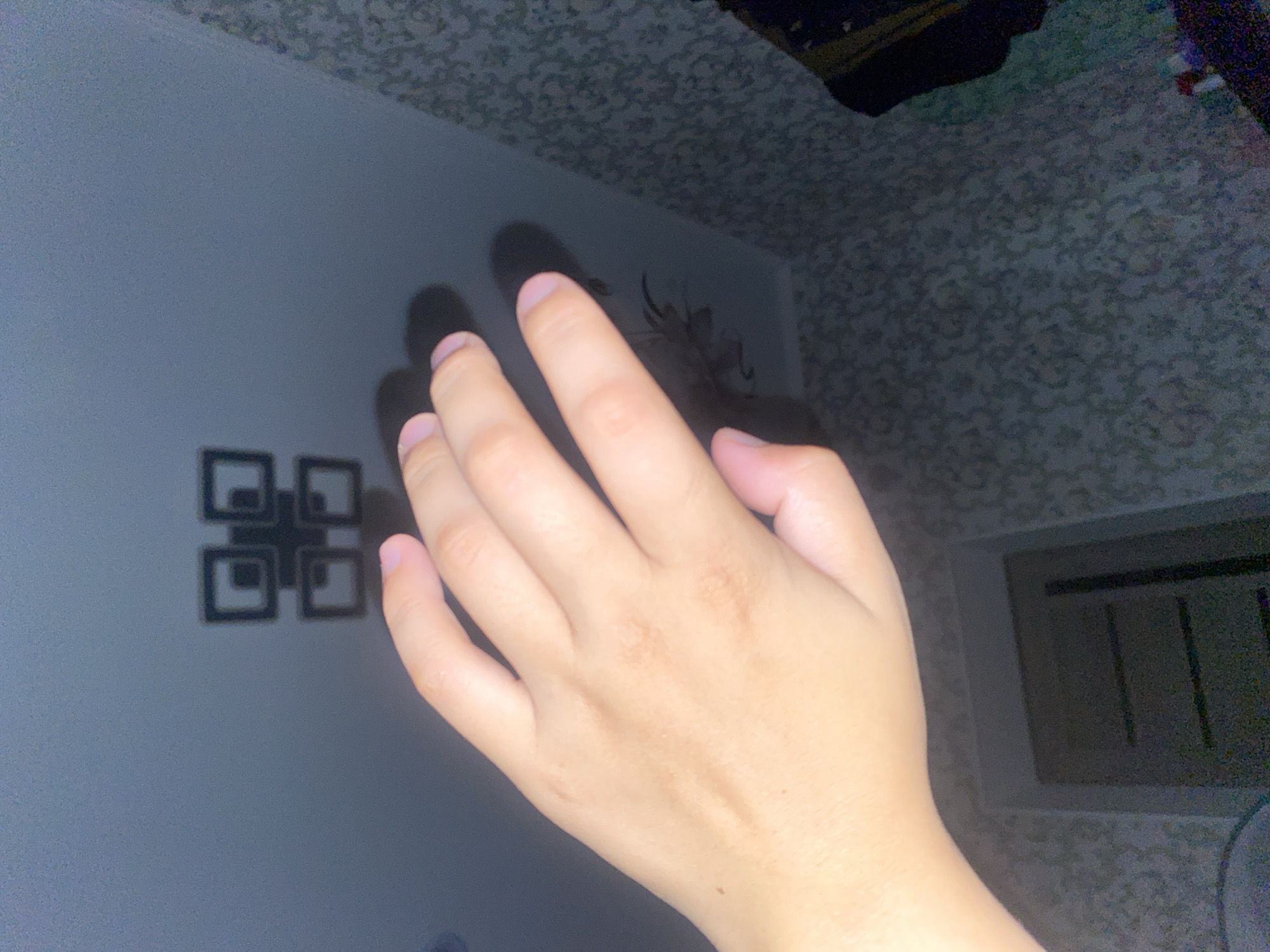 The image shows a hand with pale skin and light fingernails extended towards a ceiling. The ceiling is dark gray and features a geometric light fixture composed of four interconnected squares, forming a larger square shape. A shadow cast by the hand is visible on the ceiling, partially obscuring the light fixture. The background of the image is a wall with textured wallpaper, patterned with small, light-colored floral or abstract designs. To the right of the wall, part of a dark clothing item is visible. Below the wall and to the left, a doorway with a dark-colored door is present. The image does not provide any information about the city of Massy, Kyrgyzstan.
