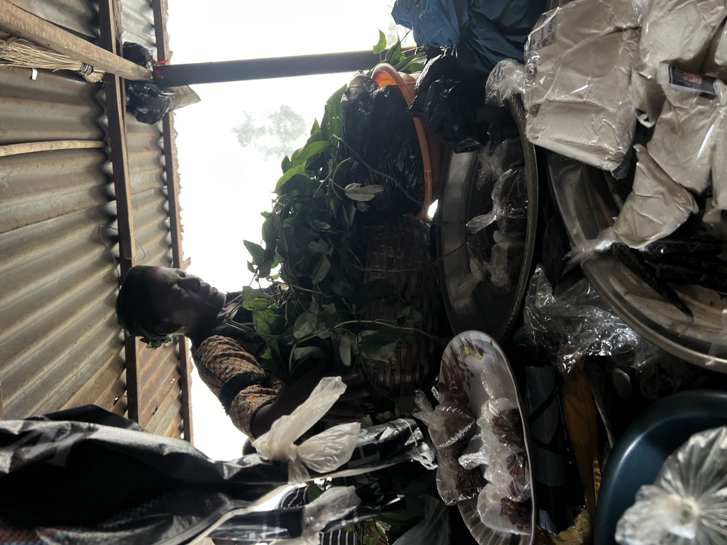 This image captures a close-up scene inside a market stall or small shop in Nkpor, Nigeria, during daylight hours. The primary subject is a woman, positioned on the left side of the frame, looking upward and to her right. She has dark skin, wears a dark headscarf, and a patterned long-sleeved shirt with shades of orange, brown, and black. Her expression is calm and neutral, suggesting she is observing her surroundings or attending to her stall.
The setting appears to be a rustic structure, with a wall or roof section made of corrugated metal sheets visible on the left, supported by wooden beams. A broom is propped against the corrugated wall in the upper left corner. Bright, diffused light streams in from an opening above, indicating it is daytime.
The stall is filled with various goods, giving the impression of a busy commercial environment. A large, dark basket overflowing with fresh green leafy plants is a prominent feature. Surrounding this, and scattered throughout the right side of the image, are numerous items displayed on round metal trays or platters. Many products are packaged in clear plastic bags, containing dark, possibly dried, goods such as herbs, spices, or food items. Larger white plastic bags, likely holding flour or powdered substances, are also visible, some with small, dark text on white labels that are too indistinct to read. Black plastic bags are used for storage or as containers. A decorative white plate with a red and yellow floral pattern and a blue plastic bowl are also part of the display. The overall scene suggests a vibrant local market stall.