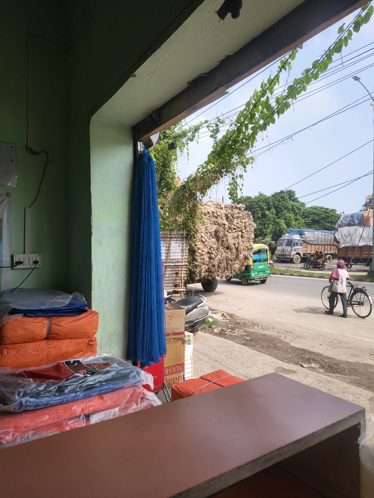 The image depicts a street scene in Purnia, India. In the foreground, a storefront is visible with stacks of bundled orange and blue tarpaulins wrapped in plastic. A large bundle of blue netting hangs vertically to the right. The storefront has a green-painted wall and a wooden beam overhead. Outside the storefront, a busy street with traffic is visible. A large truck loaded with jute sacks is a prominent feature, alongside a green auto-rickshaw. In the background, other trucks, trees, and utility poles can be seen. A woman is depicted pushing a bicycle on the side of the road, and a motorcycle is parked on the left side of the street. The sky is clear and blue.