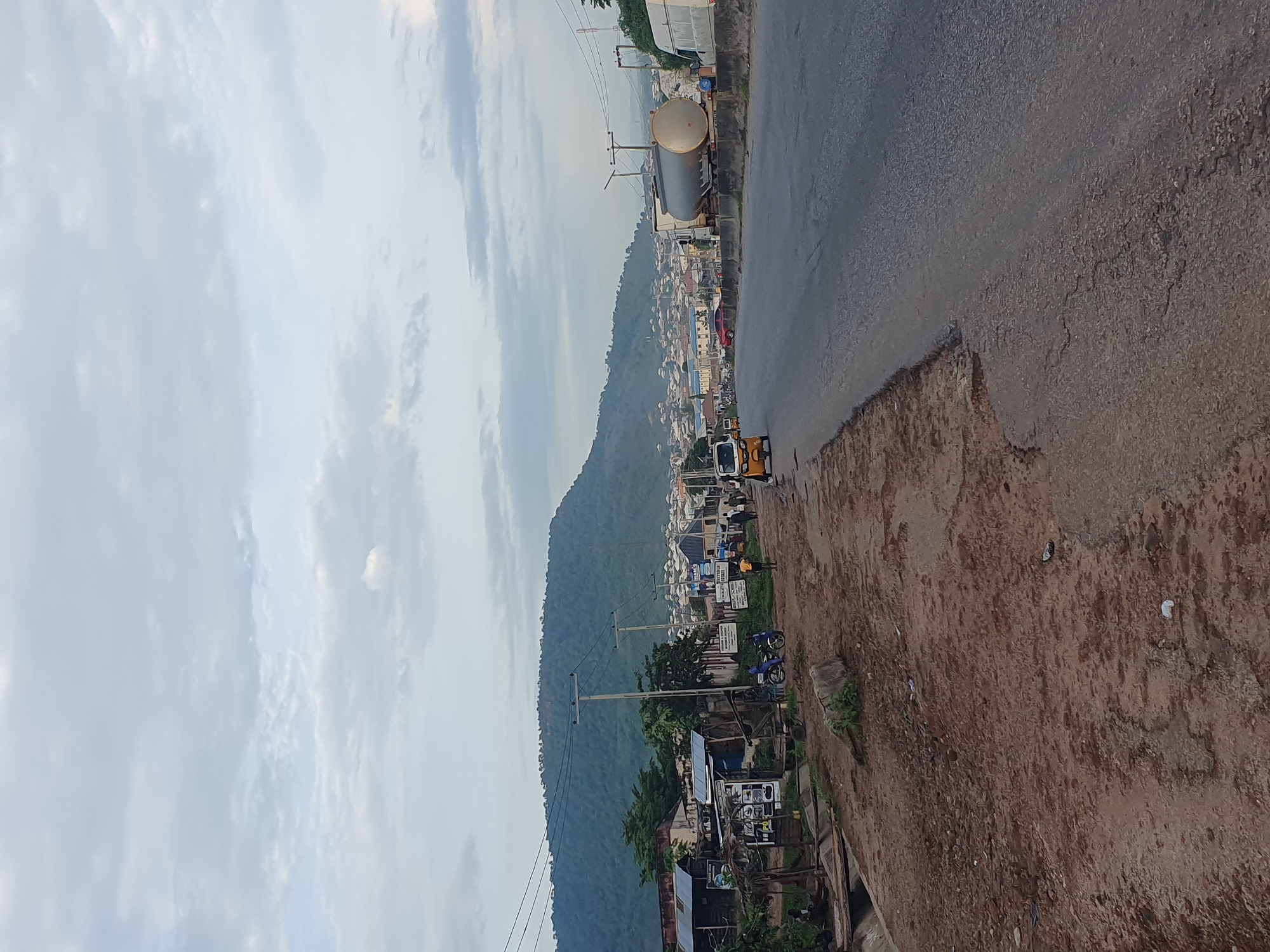 The image depicts a roadside scene in Lokoja, Nigeria, likely in the early morning or late afternoon, given the soft lighting. A paved road runs diagonally from the lower right to the middle distance, bordered on the right by a concrete barrier and on the left by a dusty, unpaved shoulder. Along the left shoulder are several low-rise buildings, some appearing to be shops or market stalls, with corrugated metal roofs. Power lines are visible overhead, supported by utility poles.
In the mid-ground, a tanker truck with a large cylindrical tank is parked on the right side of the road, with a white van behind it. A yellow tricycle, commonly known as a "Keke Napep" or "Marwa," is seen on the left side of the paved road, moving away from the viewer. Several individuals are present, including a person near a motorcycle on the left, and a few figures walking or standing along the roadside in the middle distance. The background is dominated by a large, tree-covered hill or mountain, with a settlement of houses visible cascading down its lower slopes. The sky is overcast with light, diffused clouds. The overall impression is of a busy, functional road on the outskirts of a town, with signs of commerce and daily life.