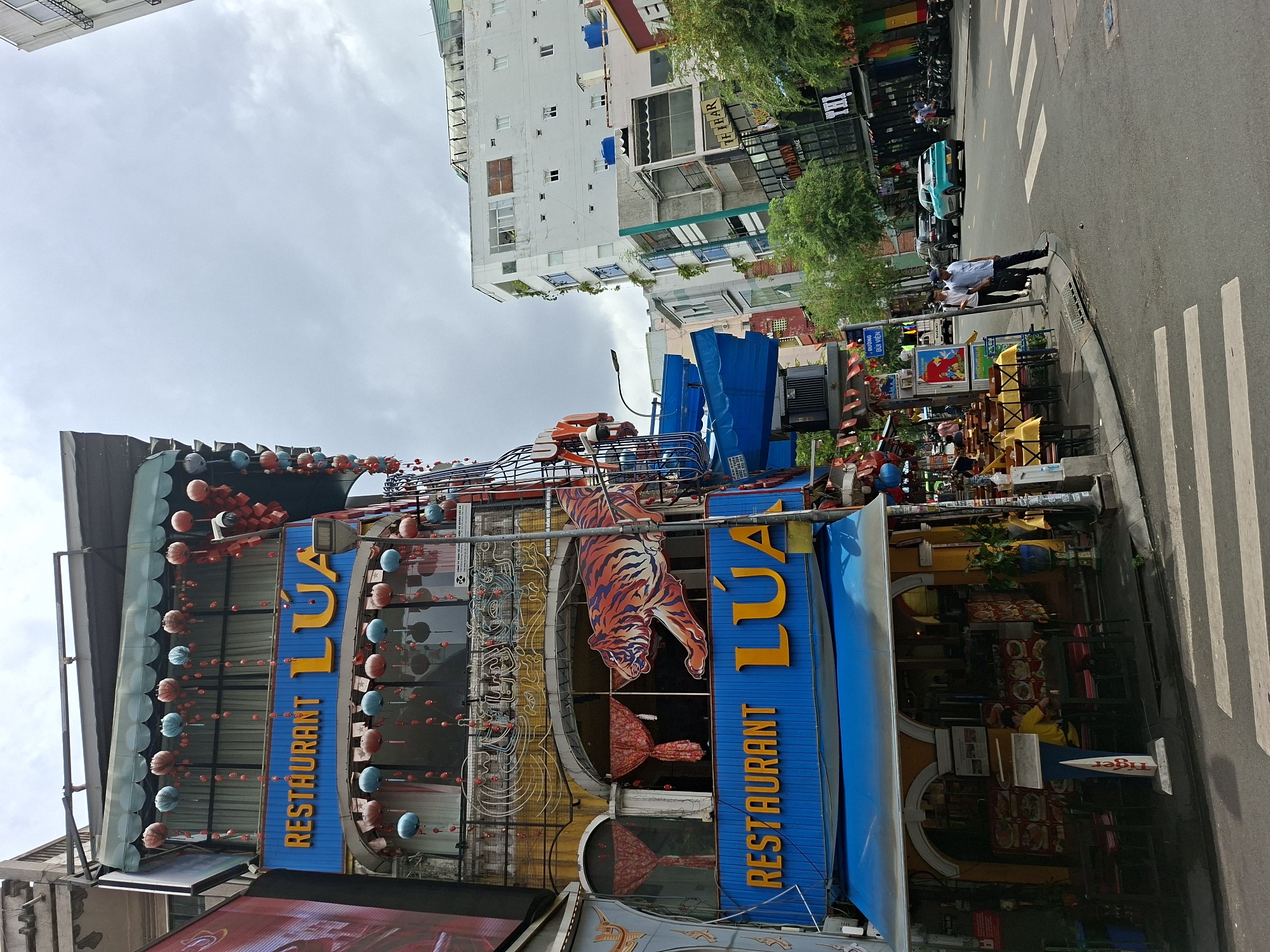 A medium shot captures a street scene in Thủ Đức, Vietnam, on a cloudy day. The prominent building on the left is a multi-story establishment with a blue facade on its lower levels, bearing the text "RESTAURANT LUA" in yellow lettering. This building is adorned with numerous red and blue lanterns hanging from its exterior, and a large, stylized orange and black tiger graphic is displayed on the second story. The ground level of the building features an entrance with yellow awnings and visible seating areas inside, where one person is seated at a table. A street crosses in the foreground, marked with white pedestrian stripes. Across the street, several other buildings line the road, including a tall white structure and other shops with colorful signage. A turquoise car is parked on the right side of the street, and several motorcycles are visible further down. Two men are walking on the sidewalk on the right side of the street, one in a white shirt and dark pants, the other in a blue shirt and dark pants. Trees are interspersed along the sidewalk. The overall impression is a bustling urban environment with a clear focus on the "RESTAURANT LUA" building.