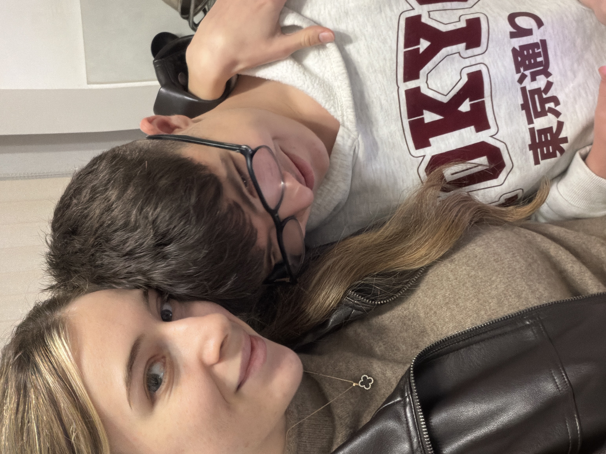 A selfie depicts a fair-skinned female with blonde hair and green eyes, lying in close proximity to a fair-skinned male with short brown hair and black-rimmed glasses. The woman is wearing a brown sweater and a black leather jacket, and a gold necklace with a four-leaf clover pendant is visible. The man is wearing a grey sweatshirt with the letters "OKY" and the Japanese characters "東京通り" in maroon. The location cannot be determined from the image. No actions or interactions can be definitively ascertained.