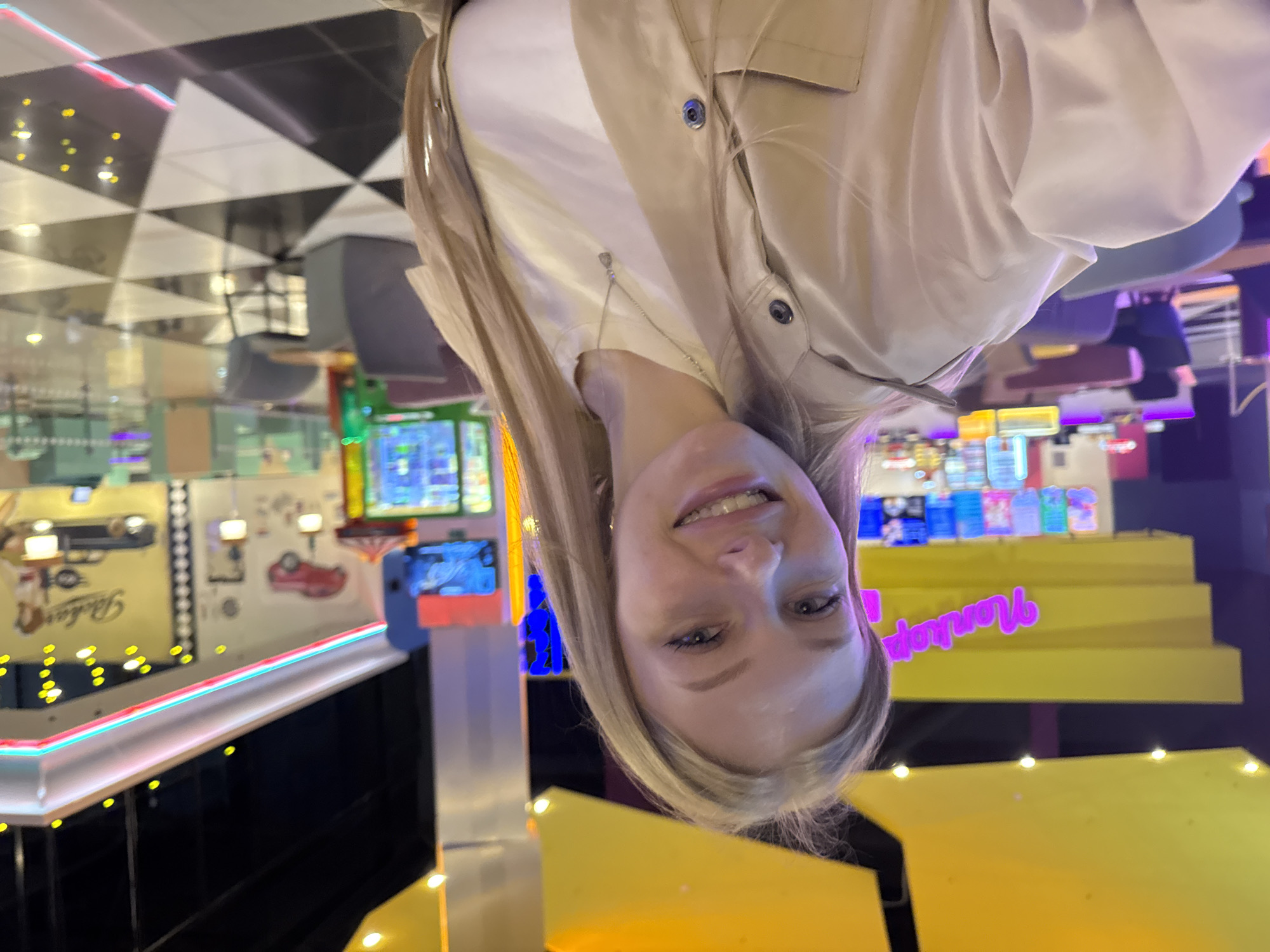 The image is of a fair-skinned person with blond hair, facing the camera. The background suggests an entertainment center, specifically a retro-themed arcade or amusement area. There are arcade games and a checkered pattern on the ceiling. There is a light-up sign that says "Vaniokop". This location is likely within the city of Perm, Russia.