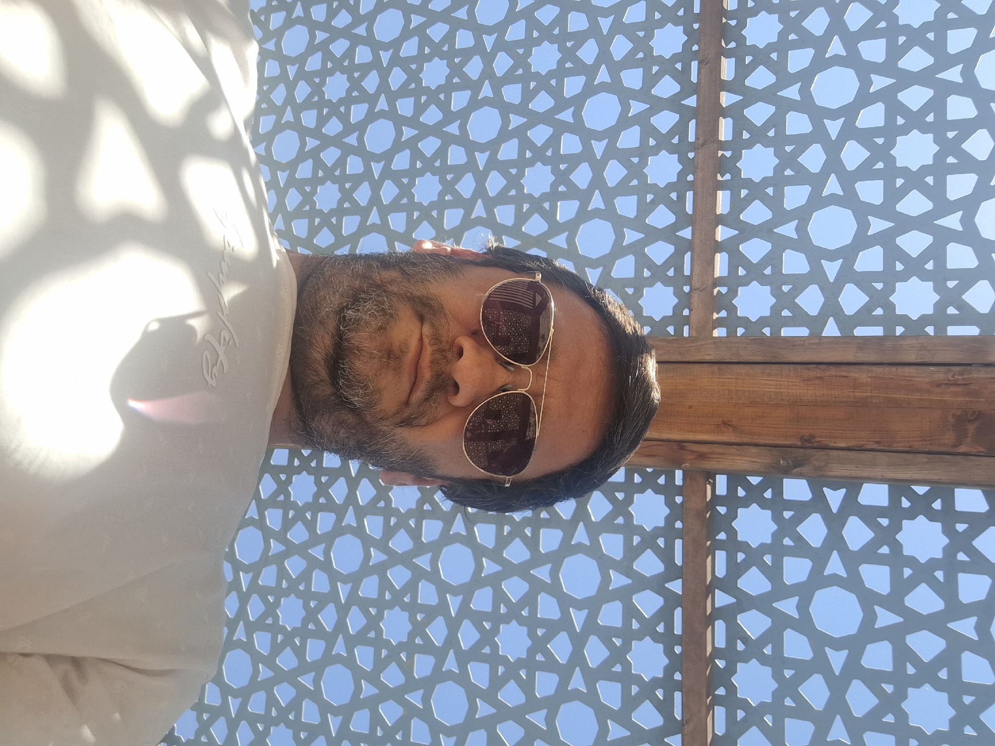 A low-angle, outdoor, waist-up selfie is taken of a man wearing aviator sunglasses and a white shirt. The man has dark hair and a beard. He is partially shaded by a wooden structure and a white patterned screen that allows sunlight to filter through. The background is a clear blue sky. The image appears to be taken in Yerocham, Israel, possibly in a park or outdoor seating area with decorative screening.