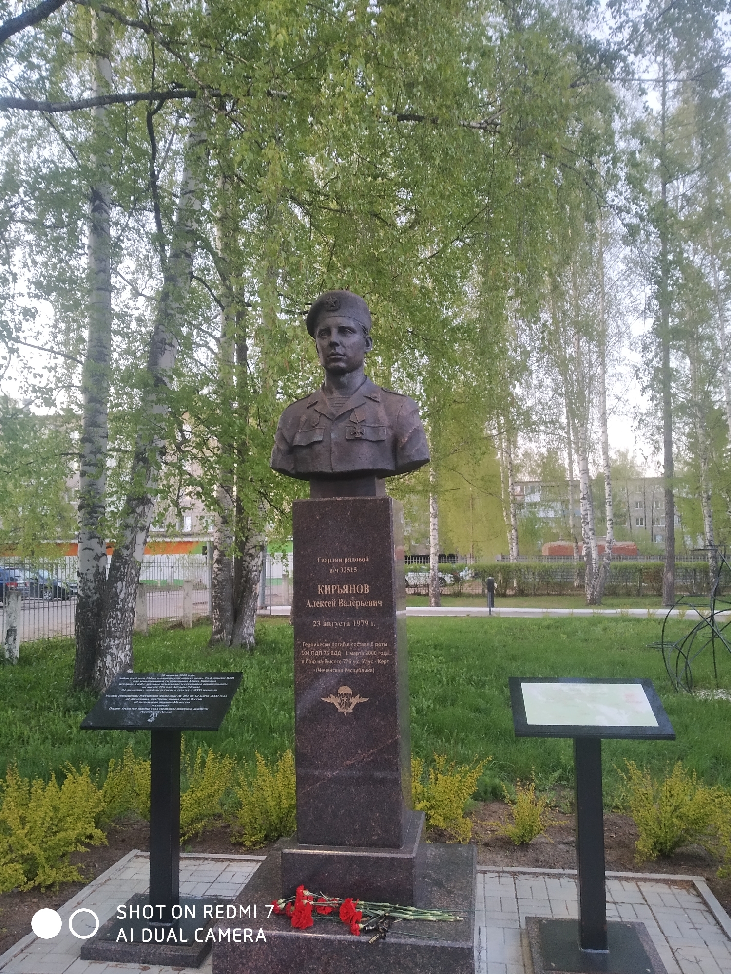 The image depicts a bronze bust of a military man with a beret in a park-like setting. The bust is mounted on a dark marble pedestal with an inscription in Russian, identifying the person as Kiryanov Alexey Valeryevich, born on August 23, 1979, and stating he heroically died as part of the 6th Company 104th Parachute Regiment 76th Airborne Division on March 1, 2000, in battle at height 776 near Ulus-Kert, Chechen Republic. There are two additional informational signs on either side of the pedestal. Red carnations are at the base of the pedestal. The surrounding environment consists of birch trees, green grass, shrubs, and a paved area around the monument. In the background, there are buildings, suggesting the monument is in an urban setting. This is in the city of Chaikovsky, Russia.
