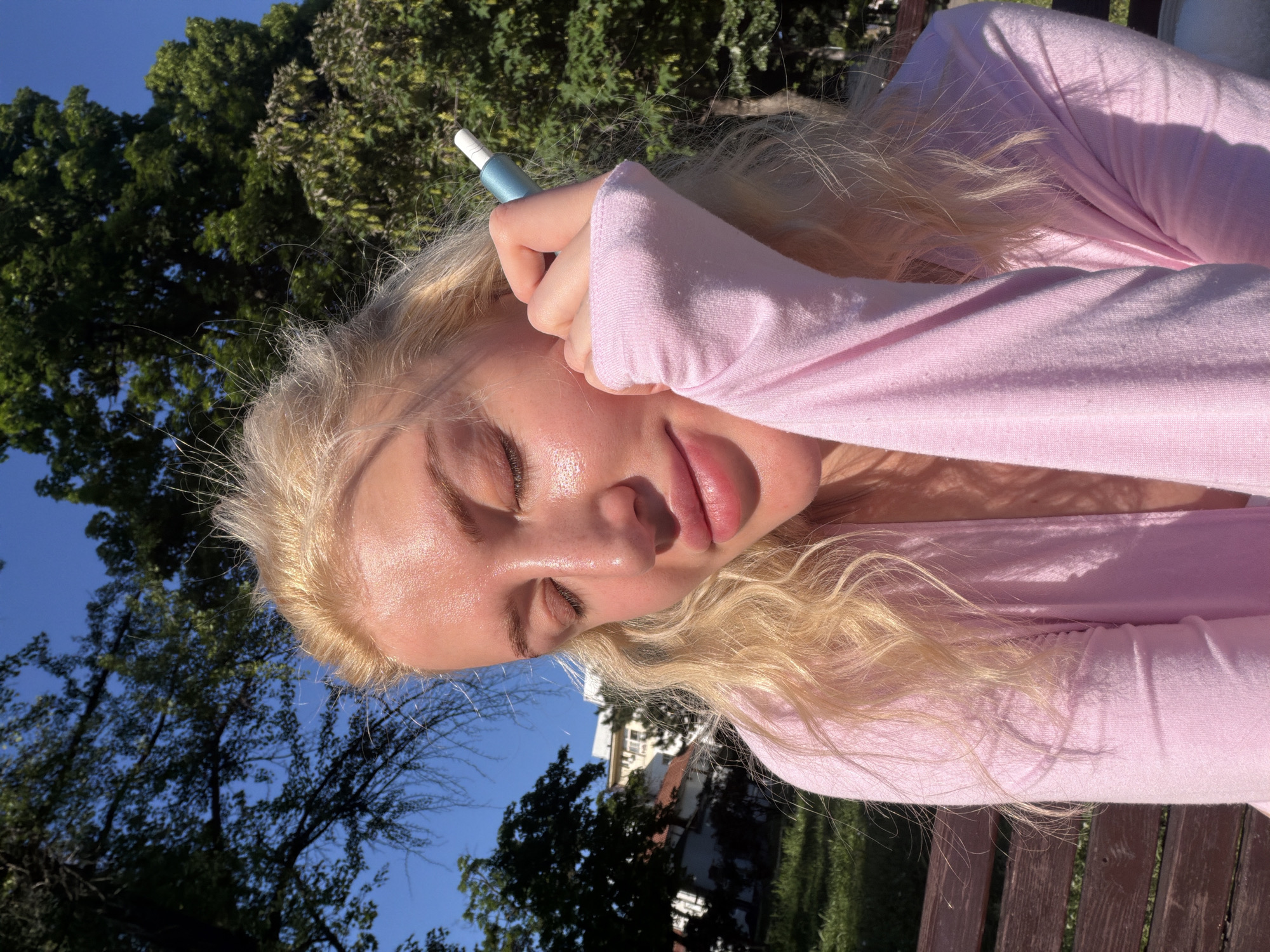 A young woman with blonde hair lies on a wooden surface outdoors, possibly a park bench. She is wearing a light pink long-sleeved top and has her eyes closed. Her hand, holding a light blue object resembling a vape pen or a similar device, is positioned near her face, resting on her forehead. The background features greenery, trees, and glimpses of buildings, suggesting an urban park or residential area. Given the context provided, it is possible that the scene is located in the Vracar Urban Municipality, Serbia. The image is taken from a low angle, looking up towards the subject.