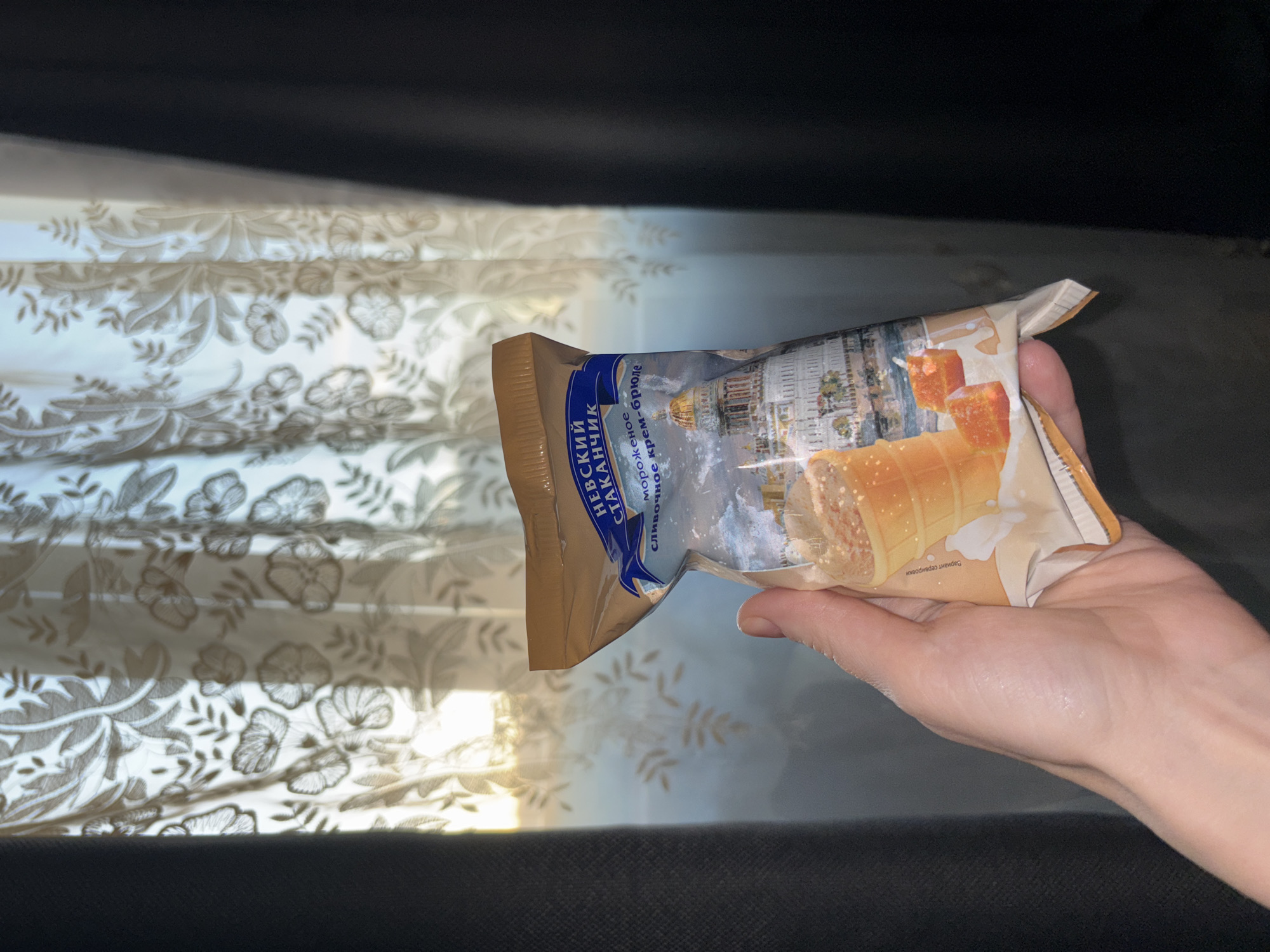A person holds an ice cream product in front of a patterned curtain. The ice cream wrapper is mostly white and brown. It features an image of a building with a golden dome. The product's name, "Невский Стаканчик", is printed in blue and white. The curtain has a floral pattern. The scene appears to be indoors, possibly near a window.