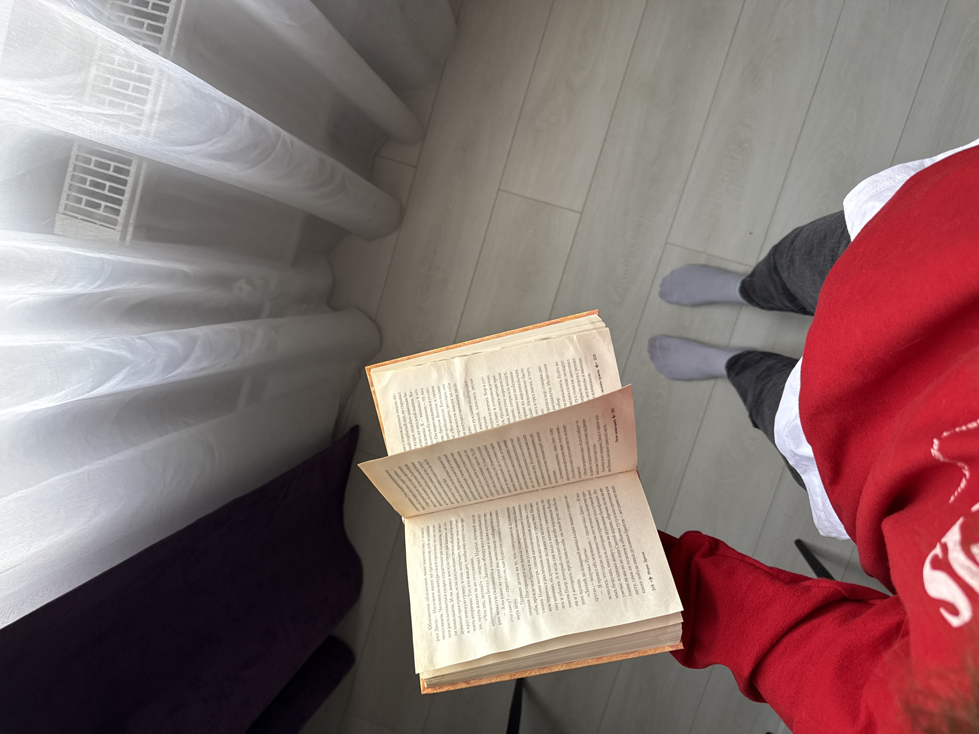 Here is a concise description of the image:
An overhead shot shows a person holding open a book with Cyrillic text. The book is held in front of a person wearing a red and white sweater, and gray pants and socks. The floor is a light, wood-grain surface. To the left is a window covered with a sheer, white curtain and a radiator. A dark purple object is visible behind the curtain.