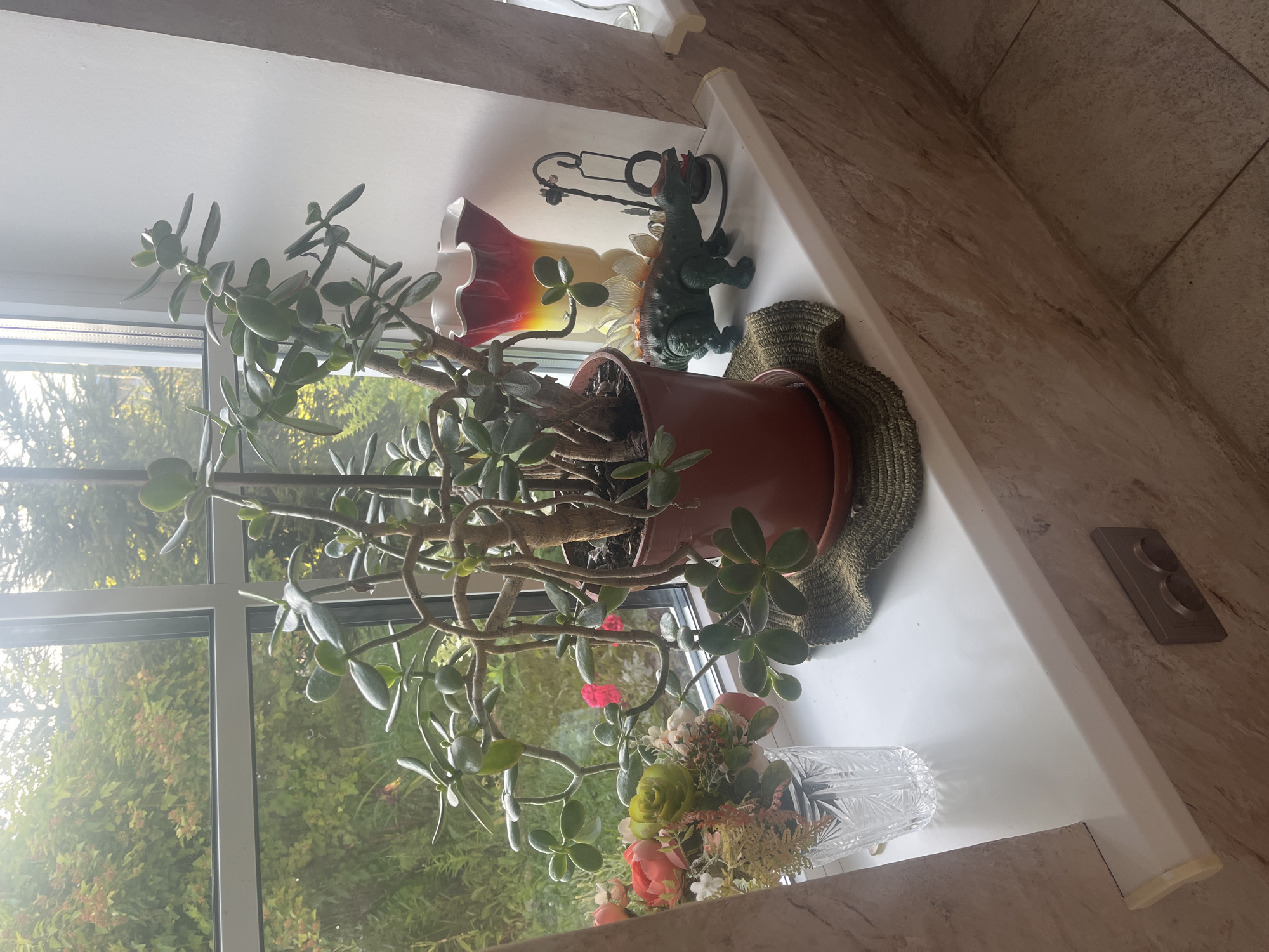 The image shows a windowsill in a domestic setting, possibly a kitchen or living room, in Vladimirovka, Russia. On the windowsill, a large jade plant in a brown pot sits prominently in the center. To the left of the jade plant, a decorative crystal vase holds a bouquet of artificial flowers, including roses and other greenery. To the right of the jade plant, a colorful vase with a red and yellow gradient is visible, alongside a dark metal decorative item. A toy dinosaur, appearing to be a stegosaurus, is positioned in front of the colorful vase. The windowsill itself is white and appears to be made of plastic. Behind the windowsill, a window looks out onto a garden with lush green foliage and hints of red flowers, suggesting an outdoor environment. The wall below the windowsill is tiled with a light-colored, marbled pattern, and a light switch with two circular buttons is mounted on the wall. The overall impression is one of indoor domestic decoration with a view of nature.