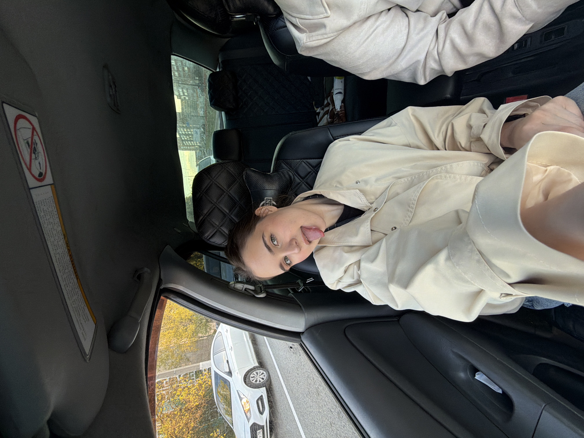 A rotated view from inside a car, showing a young adult with brown hair and green eyes sticking out their tongue. They are wearing a light tan jacket. The car interior includes black quilted seat covers, a lowered sun visor with warning labels, and a view of a white car parked on the street outside. The exterior scene suggests a street in Izhevsk, Russia, based on the car's license plate, a “18” plate indicating Izhevsk, Russia. Trees with yellow foliage are visible outside the window.