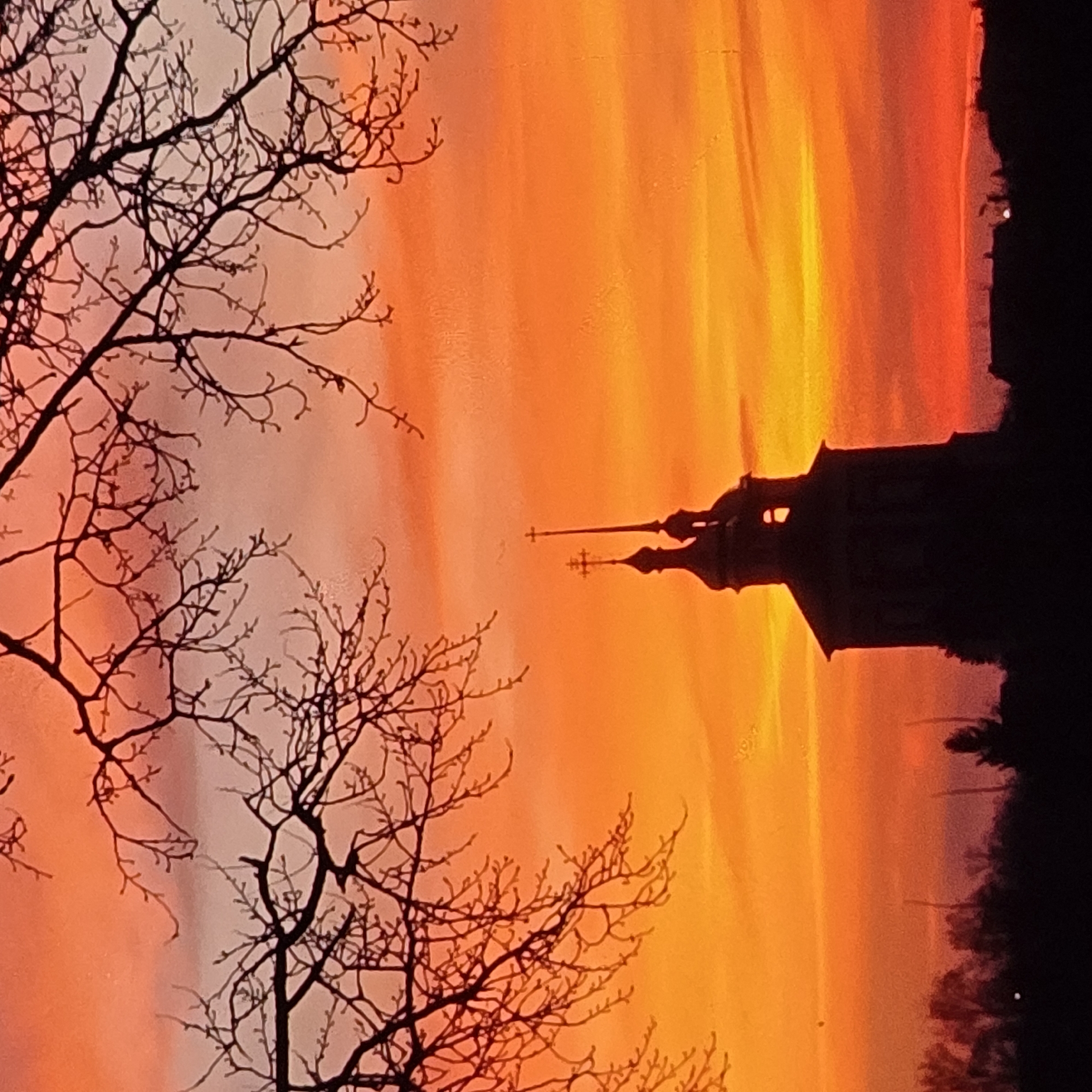 The image captures a dramatic sunset over the city of Vladimir, Russia, with the silhouette of the Dormition Cathedral (Uspensky Sobor) prominently featured. The sky is ablaze with vibrant hues of orange, red, and yellow, transitioning to a softer pink and purple near the horizon. Silhouetted against this colorful sky are the bare branches of a tree in the foreground, framing the scene.
The Dormition Cathedral, a UNESCO World Heritage site, is depicted as a dark silhouette with its distinctive onion domes and crosses. The lower portion of the image shows a dark, silhouetted treeline and structures, suggesting the urban landscape of Vladimir. The overall impression is one of natural beauty and architectural grandeur, enhanced by the dramatic lighting of the sunset. The scene is still and serene, with no people or visible activity.