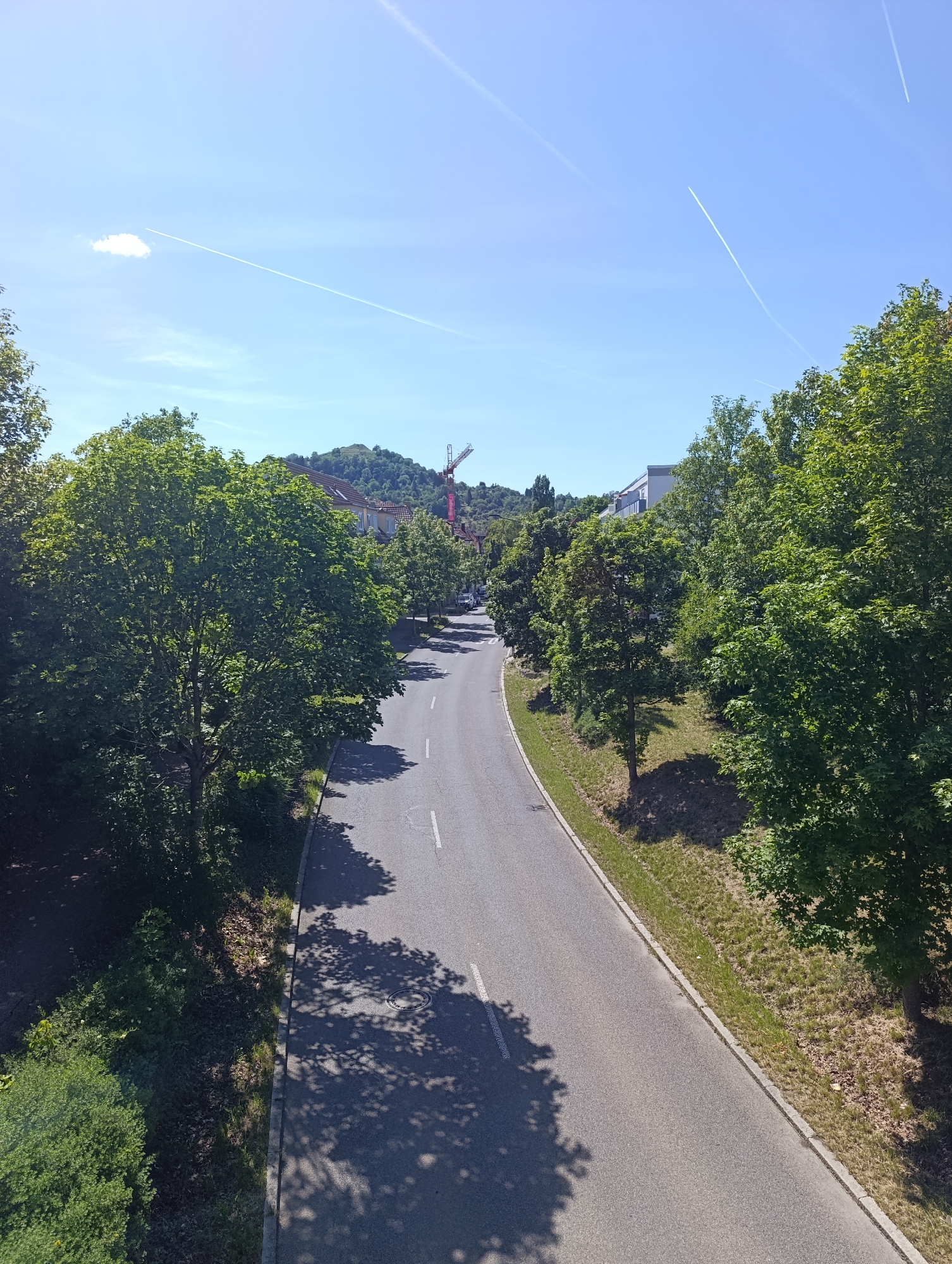 The image depicts a winding asphalt road on a clear, sunny day in Pfullingen, Germany. The sky is bright blue with several white contrails from aircraft visible. Strong sunlight casts distinct, dark shadows from trees onto the road and adjacent ground.
The road curves gently to the left from the foreground towards the midground. It features a dashed white line on its right side and a solid white line on its left, indicating lane separation. A circular metal manhole cover is visible on the right side of the road in the foreground. A concrete curb defines the edge of the road on both sides.
Dense green deciduous trees line both sides of the road, varying in height and density. Below the trees, the ground on the slopes and shoulders is covered with grass, exhibiting patches of green and drier brown areas, along with some low-lying bushes.
In the midground and background, behind the treelines, various structures are visible. On the left, several buildings, possibly residential, with different architectural styles and roof types (including one with a red-tiled roof), are partially obscured by foliage. On the right, a modern building with a light-colored facade and a flat roof is discernible. Dominating the upper right midground is a tall red-and-white tower crane, suggesting ongoing construction. A large, forested hill or mountain forms the distant background horizon.
No people, vehicles, or animals are present in the visible portion of the scene.