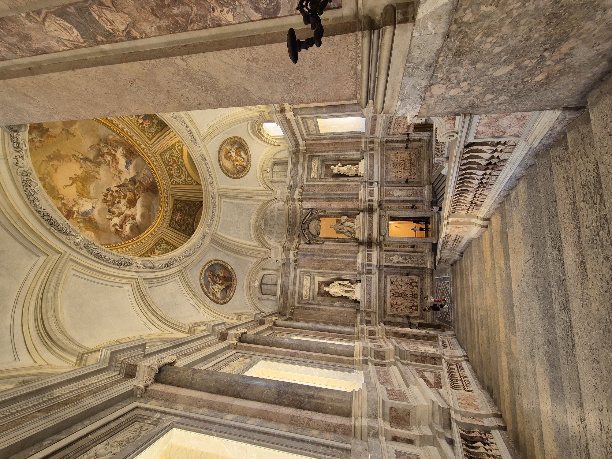 The image shows the grand staircase within the Royal Palace of Caserta, Italy. The staircase, made of stone, is wide and descends from the upper levels of the palace. It is flanked by ornate marble balustrades with intricately carved spindles. The walls of the palace are adorned with marble panels and bas-reliefs. Above, a large dome ceiling is painted with a fresco depicting celestial scenes and figures. Several statues are placed in niches along the upper levels of the staircase. Sunlight streams in from large windows, casting shadows across the steps. Two individuals, appearing to be tourists, are visible standing on a landing partway down the staircase, looking towards the ornate walls. Another person is visible further down the staircase. The scene conveys a sense of opulence and historical grandeur characteristic of the Royal Palace of Caserta.