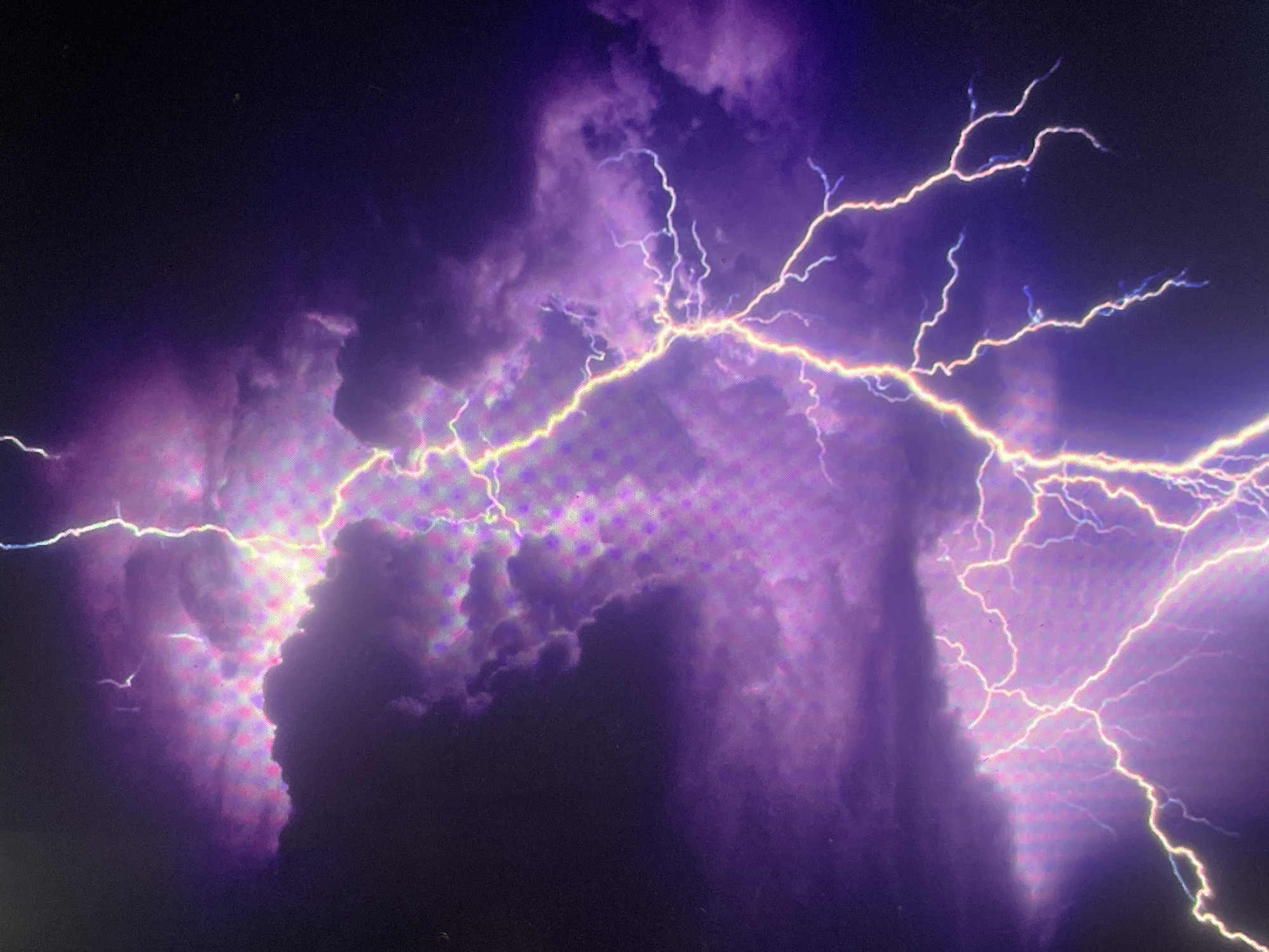 A digital image captures a dramatic lightning strike illuminating a dark, turbulent sky, identified as occurring in the city of Zelenyi Hai, Ukraine. The central and dominant feature is a vivid, multi-branched lightning bolt, primarily bright yellow and white with hints of blue, extending diagonally from the upper right quadrant towards the lower left. This bolt originates within a dense mass of dark purple and grey clouds, which are heavily illuminated from within and around by the lightning's intense light, casting vibrant purple and pink hues across the cloud formations. Smaller, thinner tendrils of lightning, appearing bluish-white, fork off the main bolt. The surrounding sky is predominantly dark black to deep indigo, with subtle gradients of purple where clouds are less dense. A noticeable grid-like pixelation pattern is visible across the entire image, particularly evident in the illuminated cloud areas, indicating a digital display artifact or a specific camera sensor characteristic. No people, man-made structures, or distinct ground features are discernible within the frame. The scene conveys a powerful atmospheric electrical discharge during what appears to be a nighttime storm.