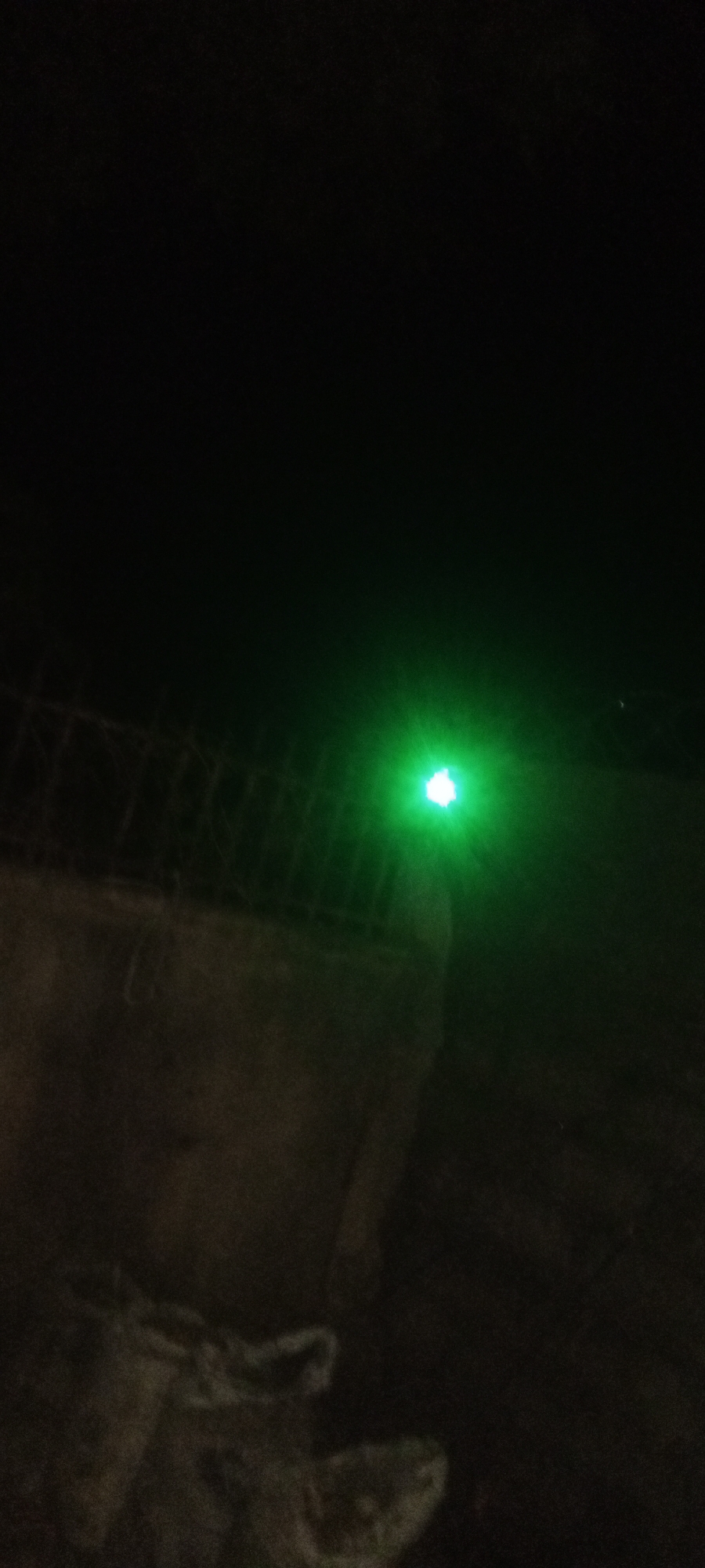 The image depicts a dark outdoor scene, likely at night. A bright green light is prominently visible against a dark background. The light appears to be shining near a wall or concrete structure topped with what seems to be a wire fence. The ground in the foreground is dark and indistinct, possibly covered in vegetation or debris. The photo does not contain any people. There is not enough information to determine the specific location within Narayi, Nigeria.