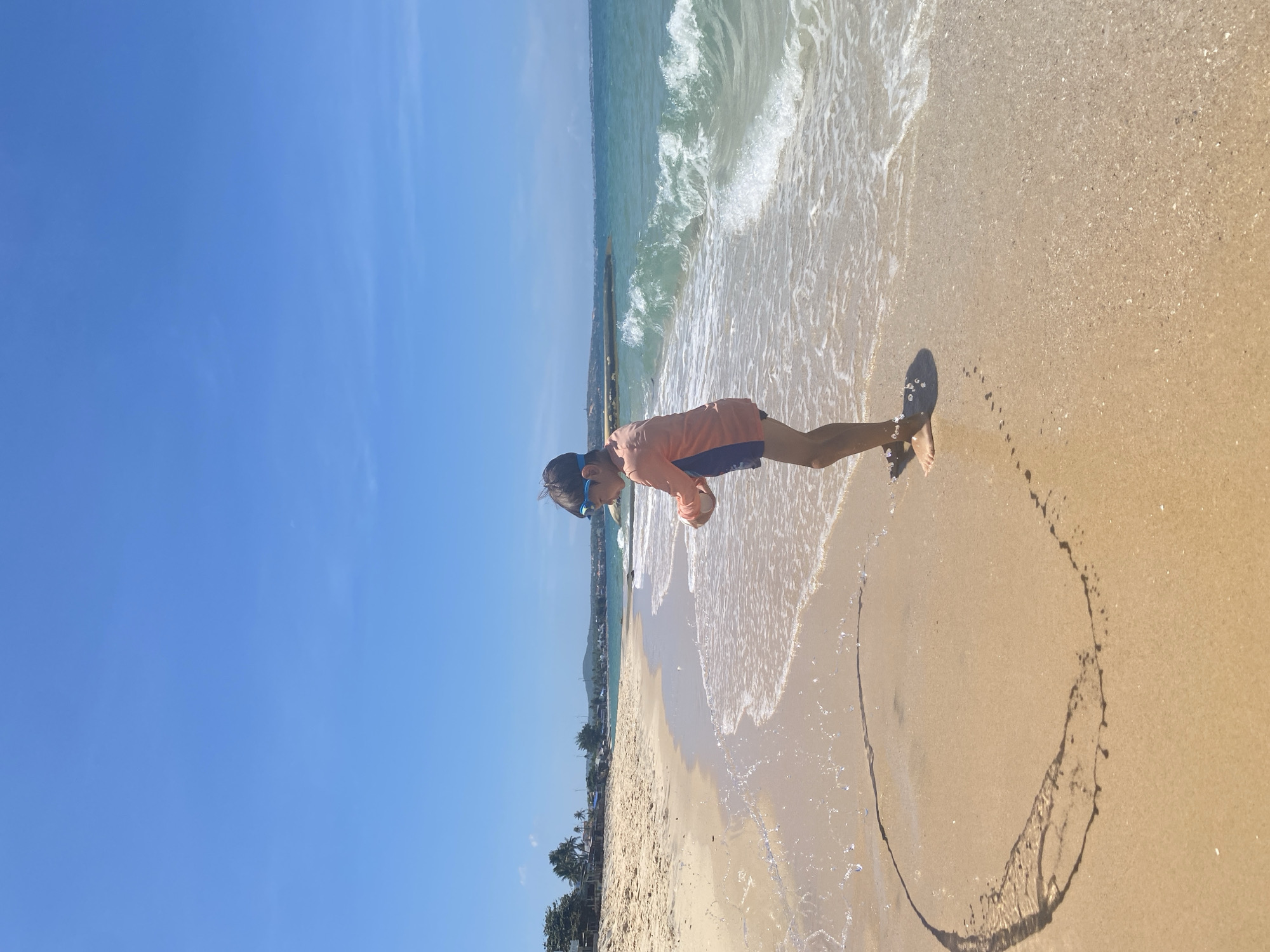 A young child wearing blue goggles and a long-sleeved orange and navy swimsuit stands on a sandy beach in Phu Thuy Ward, Vietnam, near the water's edge. The child is facing left, holding something in their hands, and appears to be interacting with the incoming waves. The ocean water is a clear turquoise with white-capped waves. The sand is light brown and wet near the water, with a circular pattern drawn in the wet sand to the child's left. In the background, there is a coastline with palm trees, buildings, and a clear blue sky. The sun is shining brightly, casting shadows on the sand.