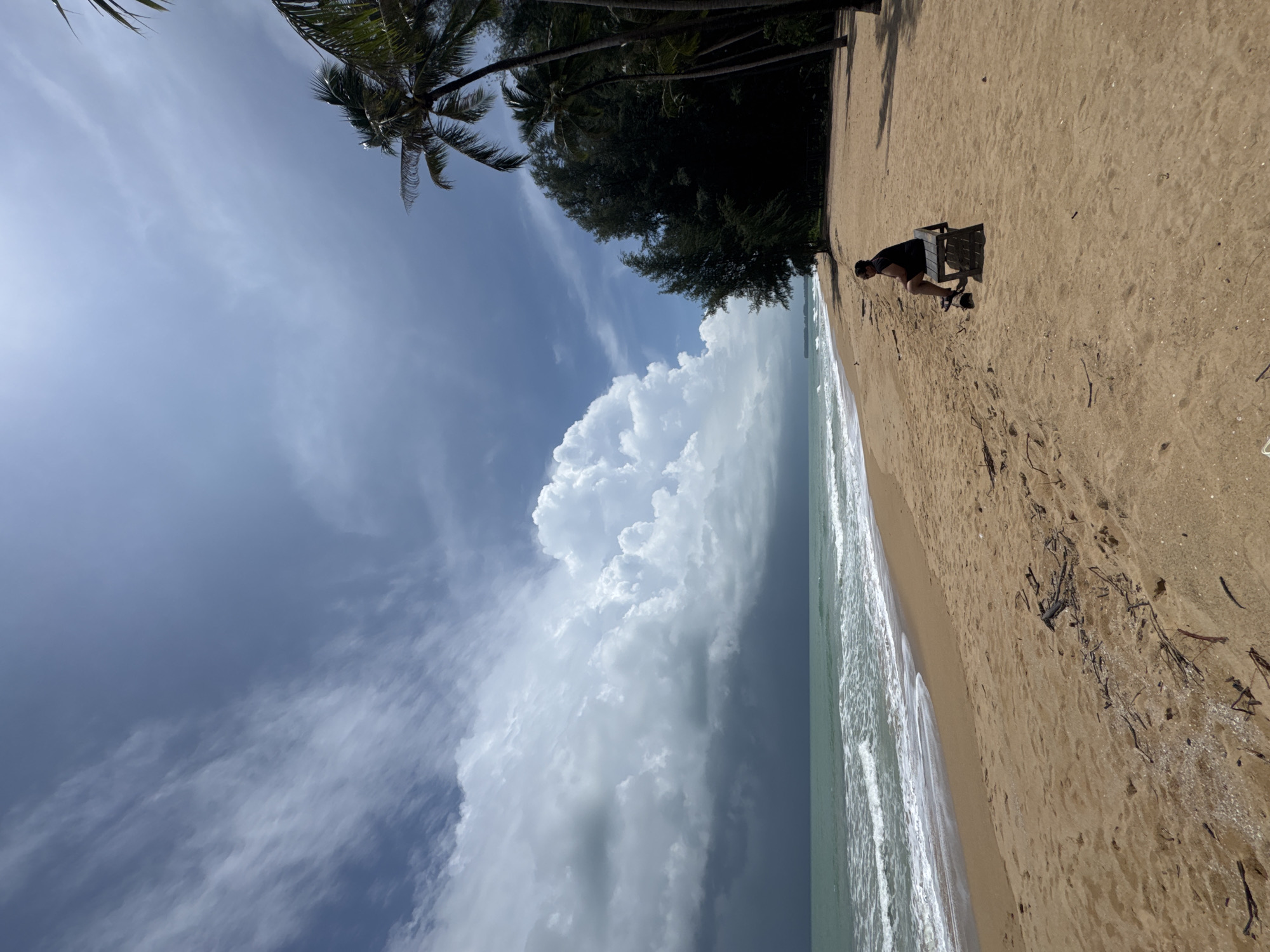 A person sits on a wooden bench on a sandy beach in Ban Khuk Khak, Thailand. The person is wearing a black t-shirt and black shorts. The beach has some debris on the sand, including driftwood. Palm trees and other foliage are visible behind the beach. The ocean is to the left of the frame, with waves rolling onto the shore. The sky is partly cloudy with dramatic storm clouds forming in the distance.