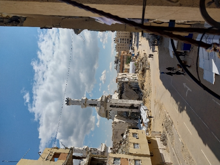 The image depicts a devastated urban landscape in Khan Yunis, Palestinian Territory, under a partly cloudy blue sky. A prominent white mosque with a single, largely intact minaret stands centrally, its main structure and dome visibly damaged and surrounded by large mounds of rubble and debris. To the left, a multi-story building is severely compromised, with its facade partially collapsed and internal floors exposed. The foreground features a paved road with a white directional arrow, traversed by several pedestrians, some in groups. Shadows are cast across the road, indicating sunlight from the left. Extensive areas of dirt and scattered building fragments flank the road and extend towards the damaged mosque. On the right, another partially visible building structure indicates the viewpoint of the photograph, with a metal balcony railing obscuring the immediate lower right corner. In the mid-distance on the right side of the road, smaller structures or stalls are visible, and more people are present in that area. Approximately 15-20 individuals, appearing to be civilians, are distributed across the scene, walking or standing amidst the destruction. Overhead wires traverse the upper mid-ground. Buildings in the distant background appear less damaged, suggesting varying degrees of destruction across the city.