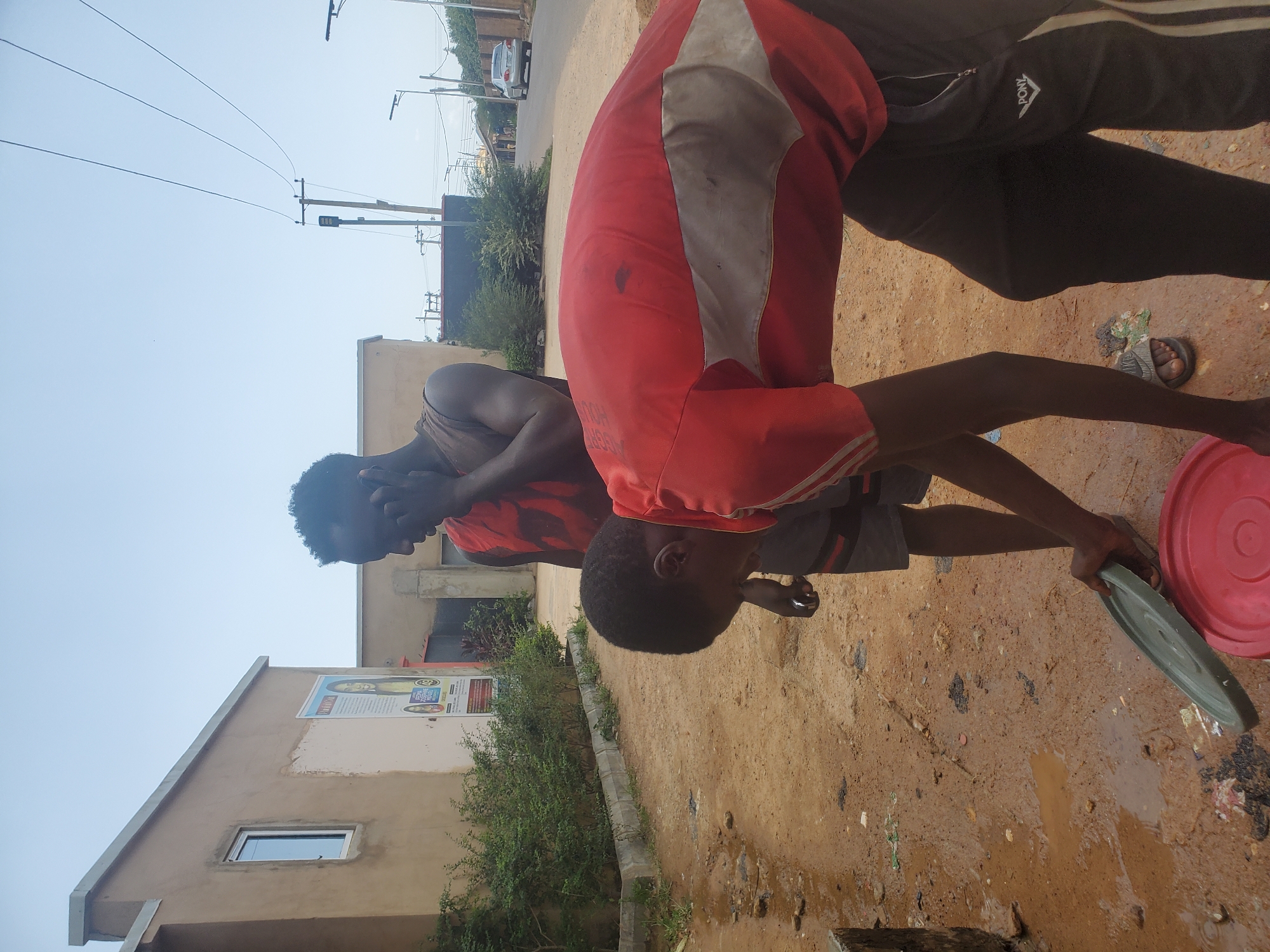 Two young men stand outside a building in Jos, Nigeria. One is washing something in a green basin over a muddy patch of ground, with a red bucket nearby. He wears a red shirt with a white stripe and dark shorts. The other man, dressed in a red and black striped top, leans his head onto his hand, watching the washing activity. The building behind them has a poster on its exterior wall, and there's a dirt road and foliage in the background. Overhead wires cross the sky.