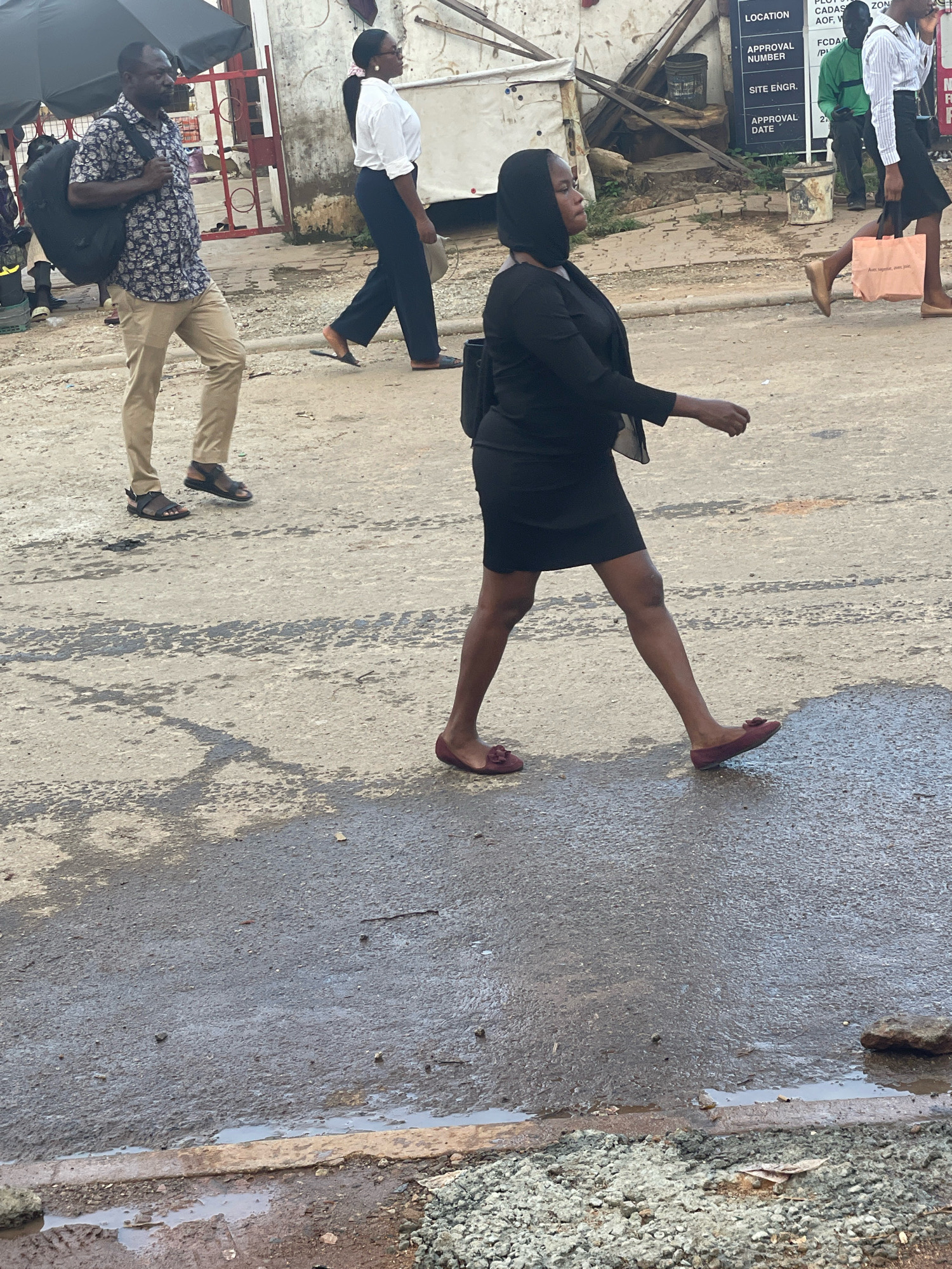 The image captures a street scene in Abuja, Nigeria, with several individuals walking on a wet, asphalt road. In the foreground, a woman dressed in black, including a headscarf, walks from left to right, carrying a black bag on her left shoulder and wearing red slip-on shoes. To her left, a man wearing a patterned short-sleeved shirt and beige trousers walks from right to left, carrying a large black backpack on his shoulders and wearing dark sandals. Behind him, a woman in a white blouse and dark trousers walks away from the camera, holding a light-colored bag. The environment shows a mixture of construction and urban elements, with debris, puddles of water on the road, and buildings in the background. A signpost with text such as "LOCATION," "APPROVAL NUMBER," and "SITE ENGR." is visible in the mid-ground, suggesting a construction or administrative area within the city. The lighting indicates an overcast day or late afternoon.