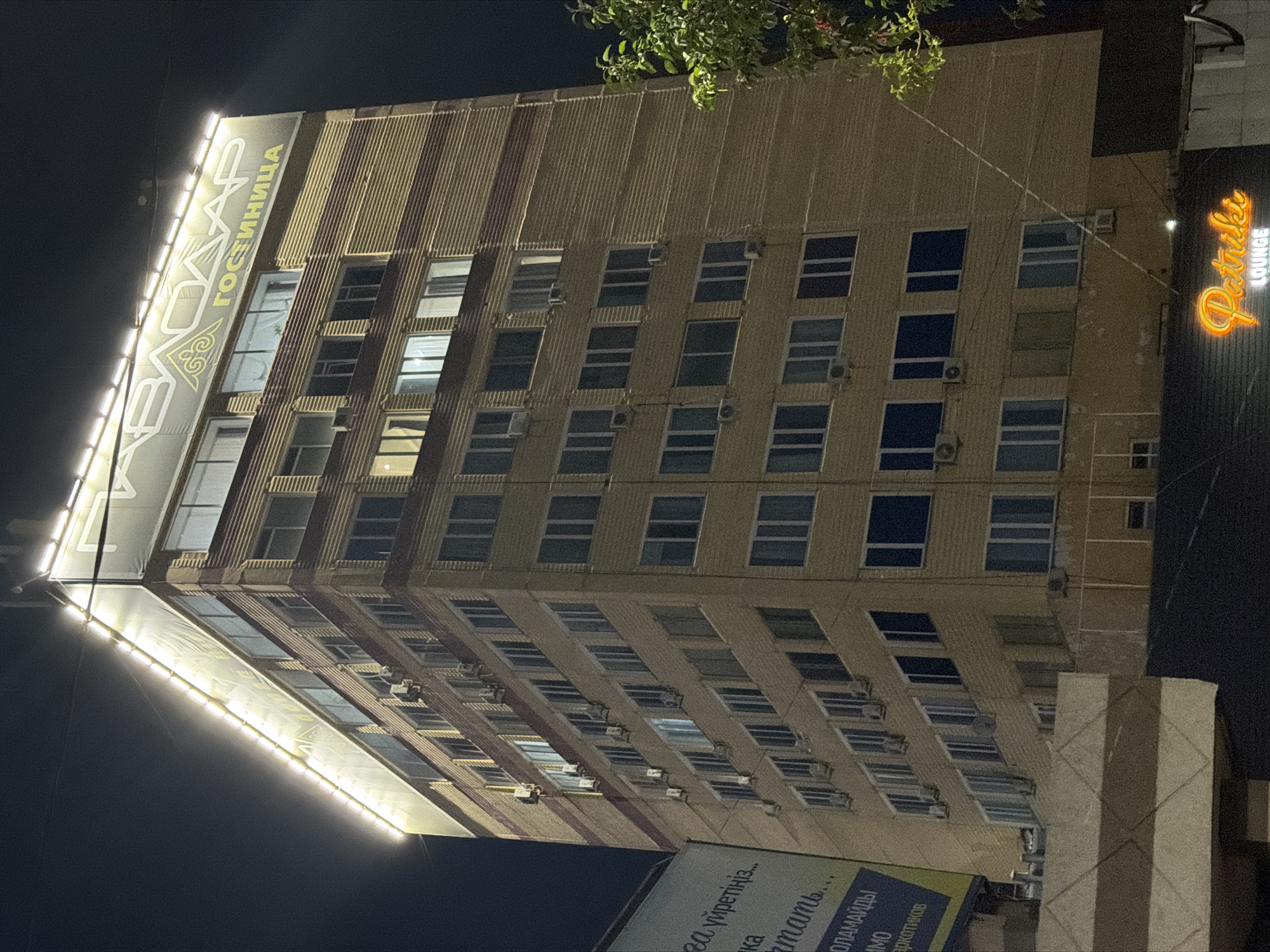 An exterior, ground-level to mid-level shot captures a multi-story, brick-clad building at night. The building is illuminated by decorative lights along its roofline and from within its numerous windows, suggesting it is a hotel or similar establishment. A large banner with yellow lettering and a decorative emblem is visible across the top portion of the building, reading "КВАЗАР ГОСТИНИЦА" in Russian, translating to "KVAZAR HOTEL". Below this, rows of windows, many with illuminated interiors and some with air conditioning units mounted outside, are visible on each floor. A neon sign with the word "Patriki" in orange script and "LOUNGE" in smaller white letters is mounted on a darker structure in the foreground, at the base of the building. A portion of a billboard with text in a Cyrillic alphabet is visible on the left side of the image. A tree with green foliage is positioned on the right side, partially obscuring the building. The overall scene depicts an urban environment at night, likely in Pavlodar, Kazakhstan, given the presence of Cyrillic script and the architectural style. There are no people visible in the image.
