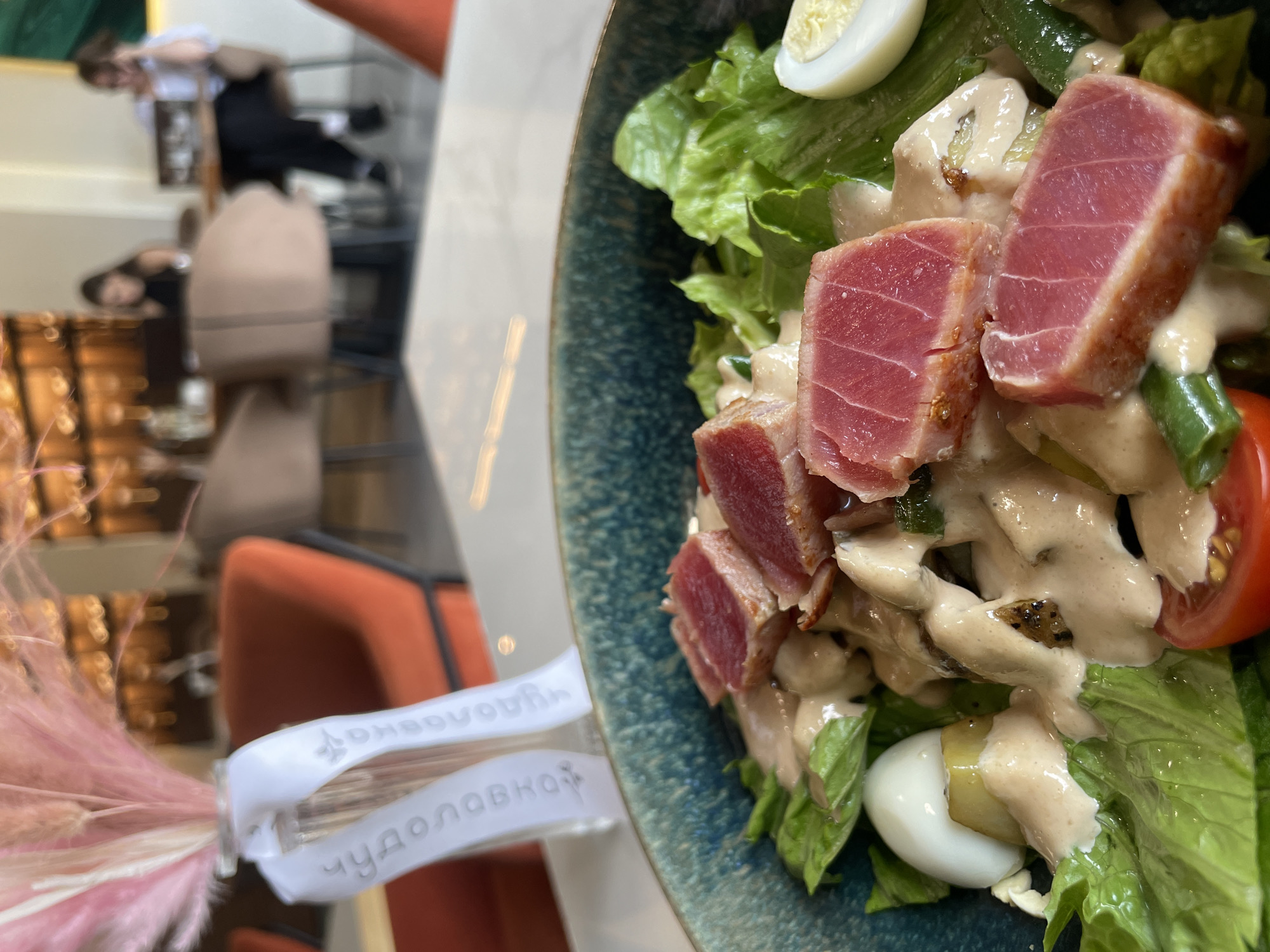 The image captures a close-up, high-angle shot of a vibrant tuna salad presented in a dark blue, textured ceramic bowl, set in what appears to be an indoor restaurant in Kazan, Russia.
The primary subject is the salad, featuring thick, seared slices of tuna with a reddish-pink interior, nestled among fresh green lettuce leaves, halved cherry tomatoes, sliced pickles, and hard-boiled quail eggs. A generous amount of creamy, light-colored dressing coats the ingredients.
In the blurred background, reflections are prominent, suggesting a highly reflective surface like a polished floor or glass table. These reflections show at least two individuals: one figure, possibly a server, dressed in a white shirt and dark trousers, stands upright. Another, less distinct reflection suggests a person seated. Reflections of orange-upholstered chairs or booths and dark tables are also visible, reinforcing the restaurant setting.
On the left side of the frame, a decorative element is visible: a bouquet of delicate pink and white dried flowers or grasses, tied with a white ribbon. Visible text on the ribbon reads "ЧУДОЛОВКА" (Chudolovka), and a partial, mirrored reflection of what might be a social media handle or brand name like "@DNBOUOVn" can also be seen.
The image focuses on the aesthetic presentation of the meal, with the blurred background creating depth and highlighting the food. The lighting suggests an indoor setting, likely during operating hours of the establishment.