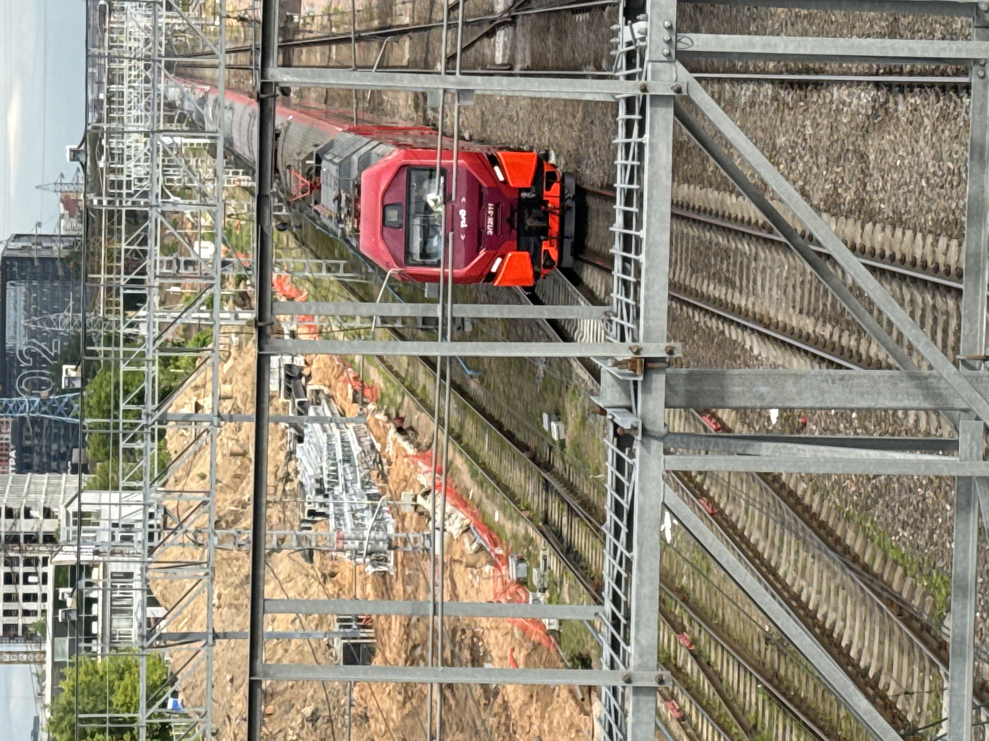 An aerial view, likely taken from an elevated structure, captures a modern red electric locomotive traveling on a multi-track railway line in an urban or industrial setting. The train, designated "ЭП2К-511" with the Russian Railways (RZD) logo visible on its front, is moving away from the viewer. A driver can be seen within the locomotive's cab.
In the foreground, a prominent metal framework, possibly part of a bridge or an overhead line support structure, partially obscures the view but also frames the railway corridor. Numerous parallel tracks with visible ballast and sleepers extend into the distance.
Adjacent to the railway lines and stretching into the background, there are areas of excavated earth and ongoing construction, marked by orange safety netting. Beyond this, a dense urban landscape featuring a mix of buildings, some under construction, forms the distant backdrop. Overhead gantries and support structures for railway electrification are present along the tracks. The scene is well-lit by daylight, indicating clear or partly cloudy weather. The setting is Moscow, Russia.