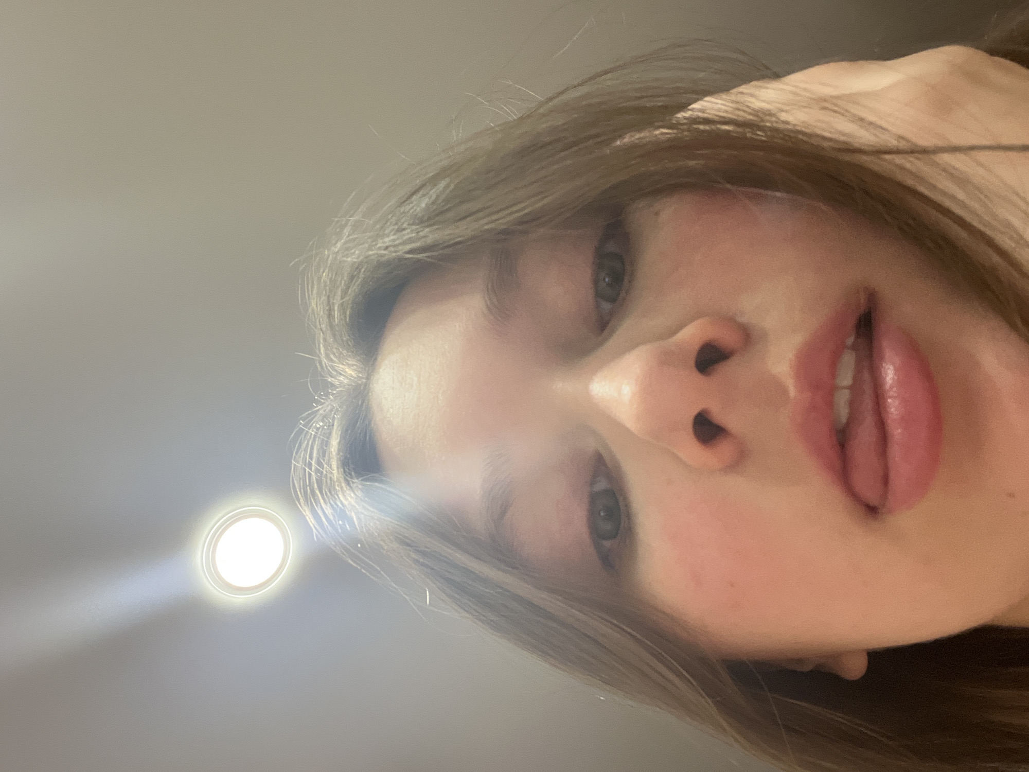 A close-up, low-angle shot of a young woman's face with light brown hair and green eyes. She is looking down at the camera. Her mouth is slightly open, and her tongue is partially visible. The lighting is uneven, with a bright light source to the left of her head, causing a glare. The background is a plain, light-colored surface, likely a ceiling. There is no indication of the city of Chaikovsky, Russia.