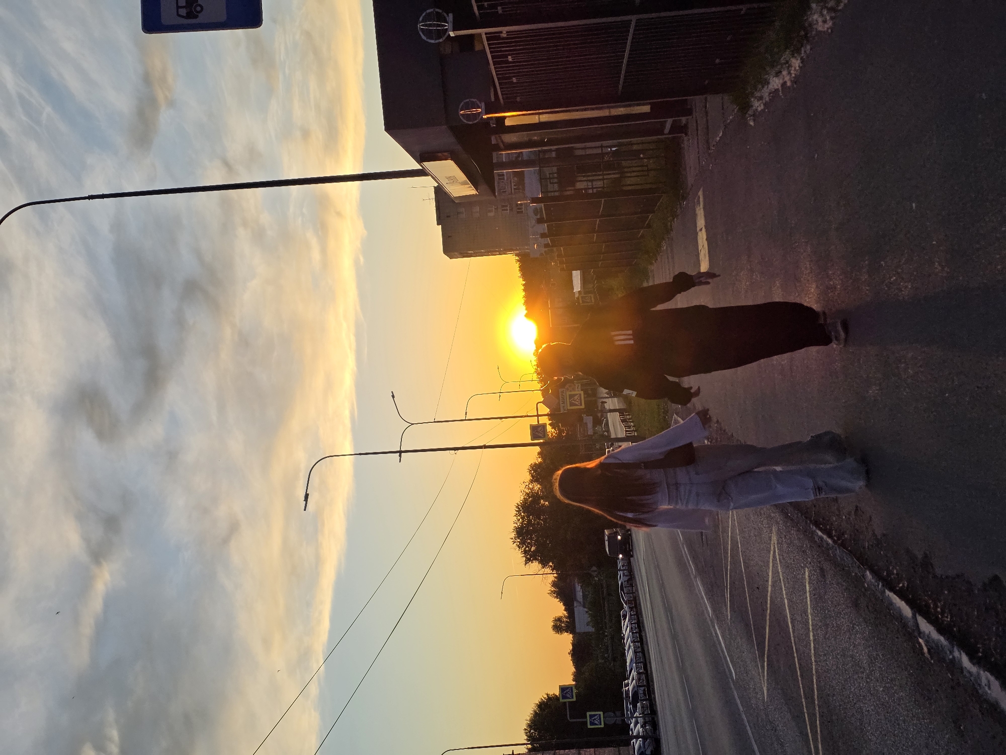 The image captures an urban street scene during a vibrant sunset in Chaikovsky, Russia. Two individuals are walking along a paved surface, likely a sidewalk or roadside. Closer to the viewer, one person wears light-colored clothing, possibly light blue jeans and a top, with long, reddish-brown hair illuminated by the sun. Further down the street, a second individual in dark clothing is partially silhouetted against the intense glare of the setting sun.
The sun, a brilliant orange and yellow orb, dominates the left side of the frame, casting a warm glow across the sky and environment. The sky exhibits a gradient of fiery orange, yellow, and soft blue, with some elongated clouds stretching across it. Along the side of the road, several cars are parked in a row. Various street furniture, including lampposts, are visible. Buildings and trees line the background beyond the street.
Notable details include a blue bus stop sign with a white bus icon in the upper left corner. Several road signs are also visible; one prominently displays the Cyrillic text "УЛ. МИРА," translating to "Peace Street." White, fluffy material, possibly poplar fluff, can be seen accumulated along the edge of the pavement. The overall atmosphere is serene, bathed in the dramatic light of dusk.