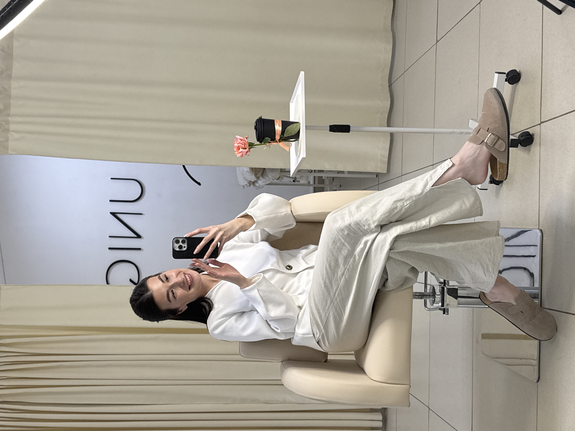 A woman is sitting in a salon chair, holding a smartphone up in her right hand, taking a selfie. She is dressed in a white linen suit and tan slide sandals. A small adjustable table holding a black pot with a peach-colored rose sits next to the chair. The background shows a curtain and a sign that reads "UNIQUE" in reverse. The location is most likely a salon in the city of Chaikovsky, Russia. The floor is tiled in a light grey color.