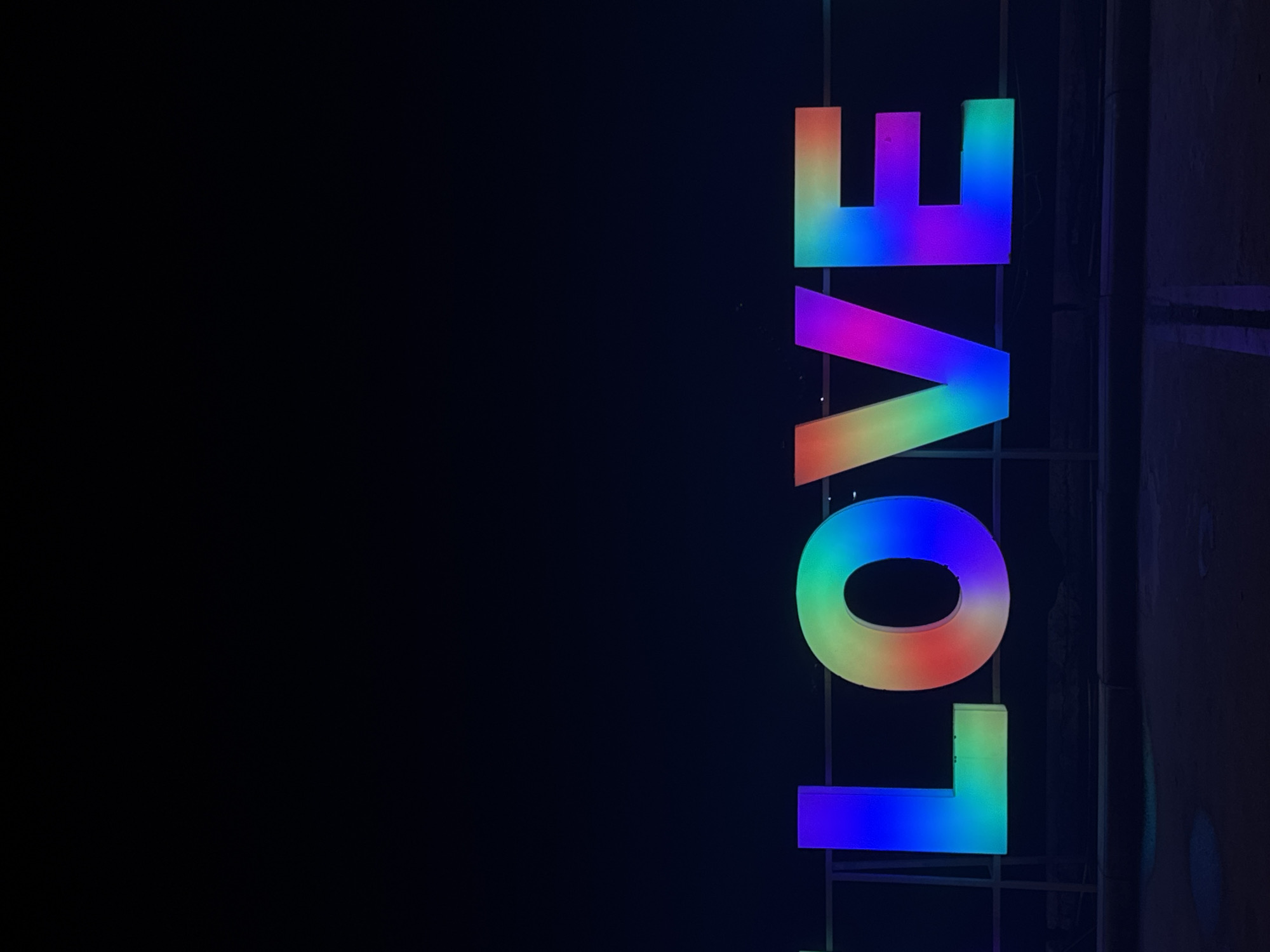 This nighttime photograph, taken in ตำบลเกาะพะงัน, Thailand, presents a large, freestanding illuminated sign spelling "LOVE" in the lower portion of the frame. Each letter is individually lit with a dynamic, multi-color gradient reminiscent of a rainbow spectrum. The 'L' exhibits a blue-to-green gradient. The 'O' displays a full chromatic sweep from red and orange on the left, transitioning through yellow, green, and blue, to purple on the right. The 'V' progresses from magenta-purple on the upper left to blue and green on the lower right. The 'E' shows a gradient from blue on the left, transitioning to green, then orange and red on the right. The sign is positioned in front of a dark, undefined background, indicative of a night sky. A dark, possibly metallic, railing or support structure is visible horizontally behind the lower section of the letters. The immediate foreground consists of a dark, paved surface, likely asphalt, with faint, lighter linear markings or stains and subtle light reflections. No individuals or discernible actions are present.