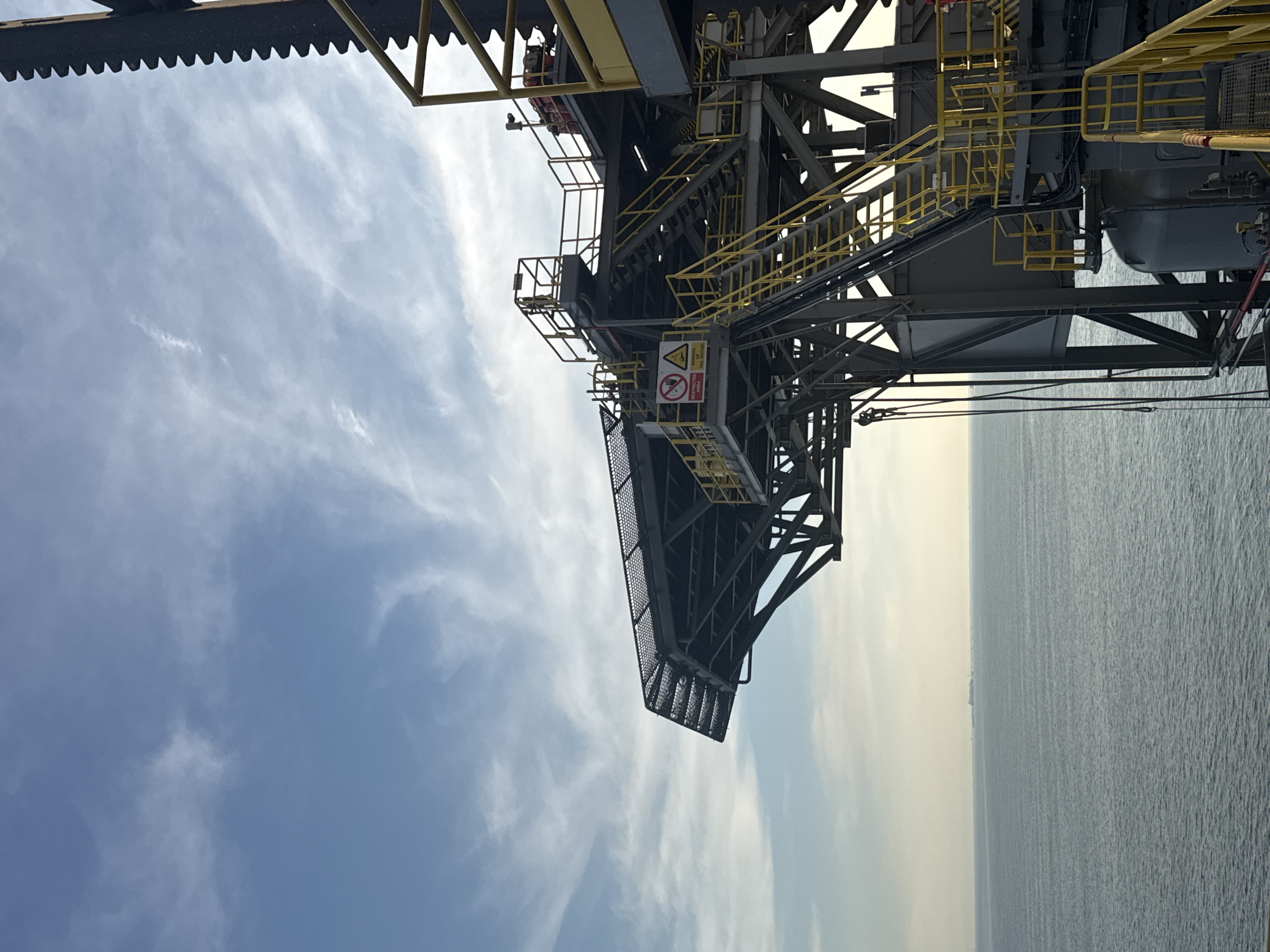 This media file displays an industrial structure, possibly an offshore oil rig or a similar large-scale offshore platform, viewed from a low angle looking upwards. The structure is characterized by its complex metal framework, featuring numerous staircases with bright yellow handrails, platforms, and what appears to be conveyor belts or chutes. The overall color palette is dominated by dark grey metal, yellow accents, and the blue of the sky.
The sky is partly cloudy with wispy white clouds scattered across a bright blue expanse, suggesting it is daytime, likely morning or afternoon. The sun is not directly visible but its light illuminates the scene, creating some highlights and shadows on the structure. Below the structure, the vast expanse of the ocean is visible, appearing calm with a subtle texture on its surface. A faint horizon line can be seen in the distance, with what might be a ship or another distant structure on it.
A notable detail is a warning sign visible on one of the platforms. It features a triangular symbol with a lightning bolt, indicating a potential electrical hazard, and a red circle with a diagonal line through a hand symbol, signifying "no entry" or "restricted area." Text below this symbol reads "RESTRICTED AREA."
The image evokes a sense of industrial scale and operational complexity. The perspective creates a feeling of looking up into a towering, intricate piece of engineering against the natural backdrop of the sky and sea. There are no visible people in the frame. The weather appears clear and pleasant for outdoor activity. The location is clearly offshore, given the presence of the ocean.