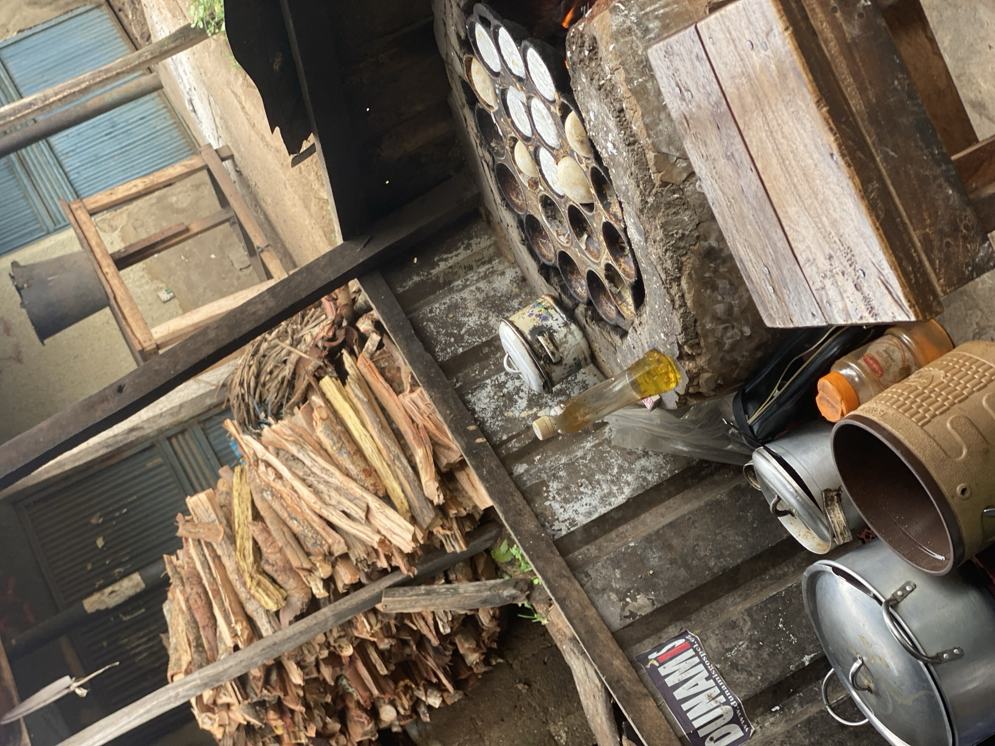 This media file depicts a rustic, semi-sheltered cooking area in Maigizo, Nigeria, likely during daytime. The primary subject is a traditional wood-fired stove, possibly made from mud or concrete, in which a large, circular, cast-iron pan with multiple individual molds is embedded. Many of these molds contain a light-colored batter, indicating active preparation of food, possibly fritters or local pancakes. Flames or embers are visible within the stove, confirming it's in use.
To the left of the stove, a substantial pile of neatly stacked split firewood serves as fuel. The ground, appearing to be dirt or worn concrete, is scattered with various objects. Several aluminum cooking pots with lids are resting on the ground, along with a small, white enamel pot. Other items include a plastic bottle holding a yellow liquid (likely cooking oil), an orange-capped container, and a black pouch.
The setting features wooden beams and planks, suggesting a basic structural framework, perhaps for a roof or shelving. In the background, corrugated metal sheets are visible, which could be part of a wall or roof, alongside a simple wooden frame. No people are visible in the image. On a dark, rectangular object in the bottom right corner, possibly a piece of paper or fabric, the word "DUNAMIS" and the website address "www.dunamisgospel.org" are clearly legible. The scene conveys a functional and utilitarian cooking environment.