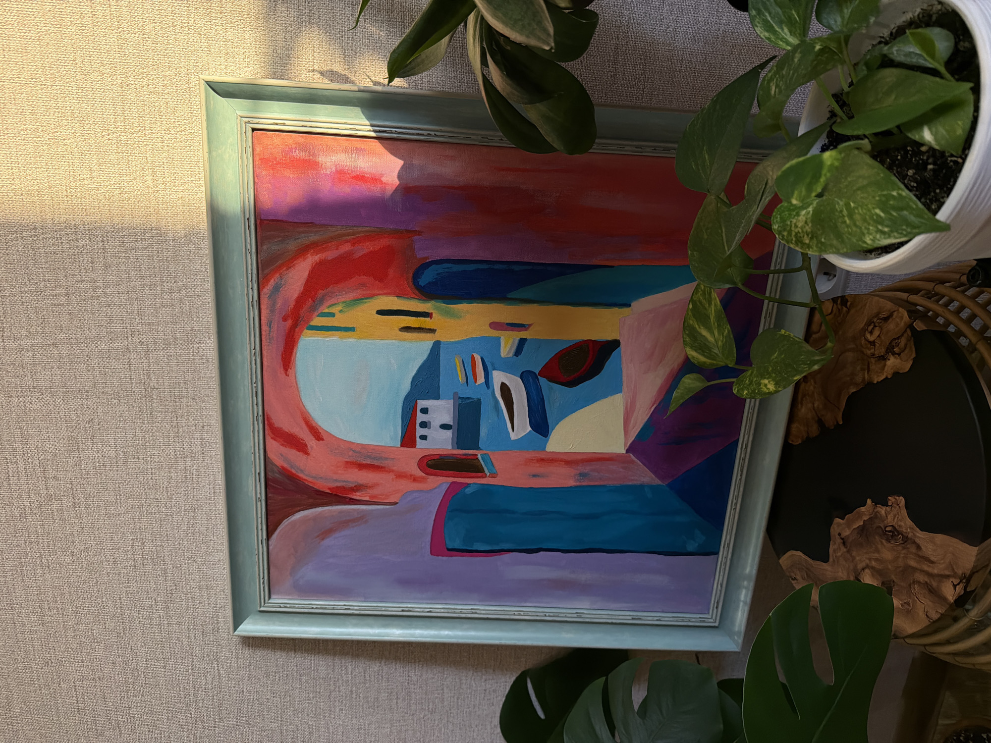 The image shows an abstract painting in a light blue frame hanging on a textured beige wall. The painting features bold strokes of red, purple, yellow, and blue. A portion of two houseplants partially obscures the painting. The plant at the top right is in a white pot, and a decorative wooden object sits next to it. Sunlight illuminates the left side of the wall. The city and exact location are unknown.