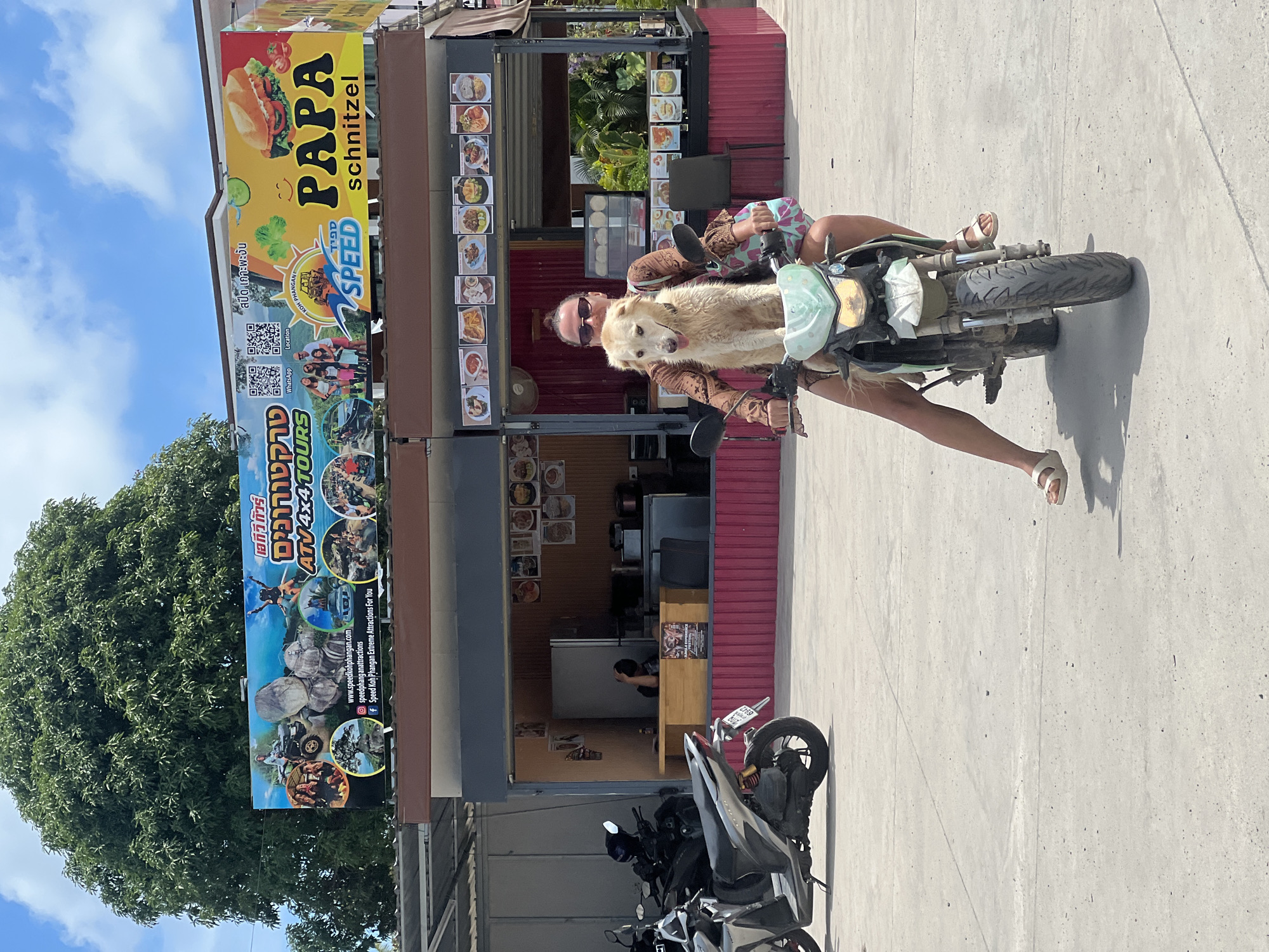 In the city of ตำบลเกาะพะงัน, Thailand, on a bright, sunny day, a woman is riding a motorcycle with a dog sitting in front of her. The woman is wearing a brown patterned dress and sunglasses, and the dog is a light-colored, medium-sized breed. The motorcycle is green and white, and the background shows a row of food stalls. The stalls have menus displayed with pictures of various dishes. Above the stalls, there are two large billboards. The left billboard advertises ATV and 4x4 tours, featuring images of ATVs, rocks, and people. The right billboard is yellow and features the text "PAPA schnitzel" and an image of a sandwich, along with a smiley face and some vegetables. In the distance, a large green tree is visible, and the sky is blue with scattered white clouds. There are also other motorcycles parked in front of one of the stalls. The ground is paved with concrete.