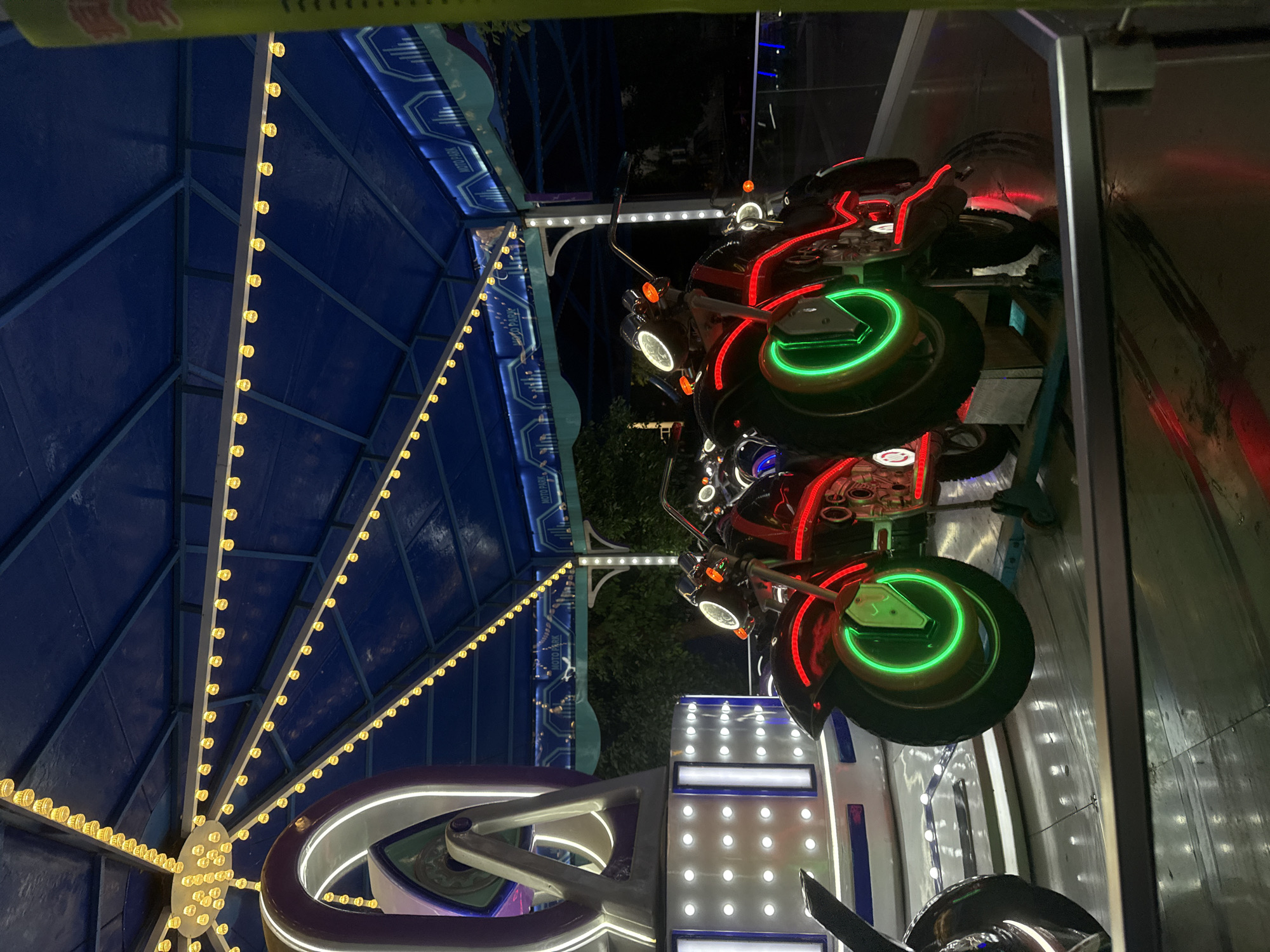 The image shows a close-up of a motorcycle-themed amusement ride, likely located at a fair or amusement park in Yiwu, China. The ride features several brightly lit motorcycles, some with neon green and red accents on their wheels and frames. The motorcycles are positioned on a platform with flashing white lights. Above, a blue canopy is adorned with strings of yellow lights. In the background, a sign reads "MOTO PARK" with stylized graphics. The overall scene is dimly lit, with the neon lights of the ride creating a vibrant and festive atmosphere. The image appears to be taken at night, with the darkness of the surroundings highlighting the illuminated elements of the attraction.
