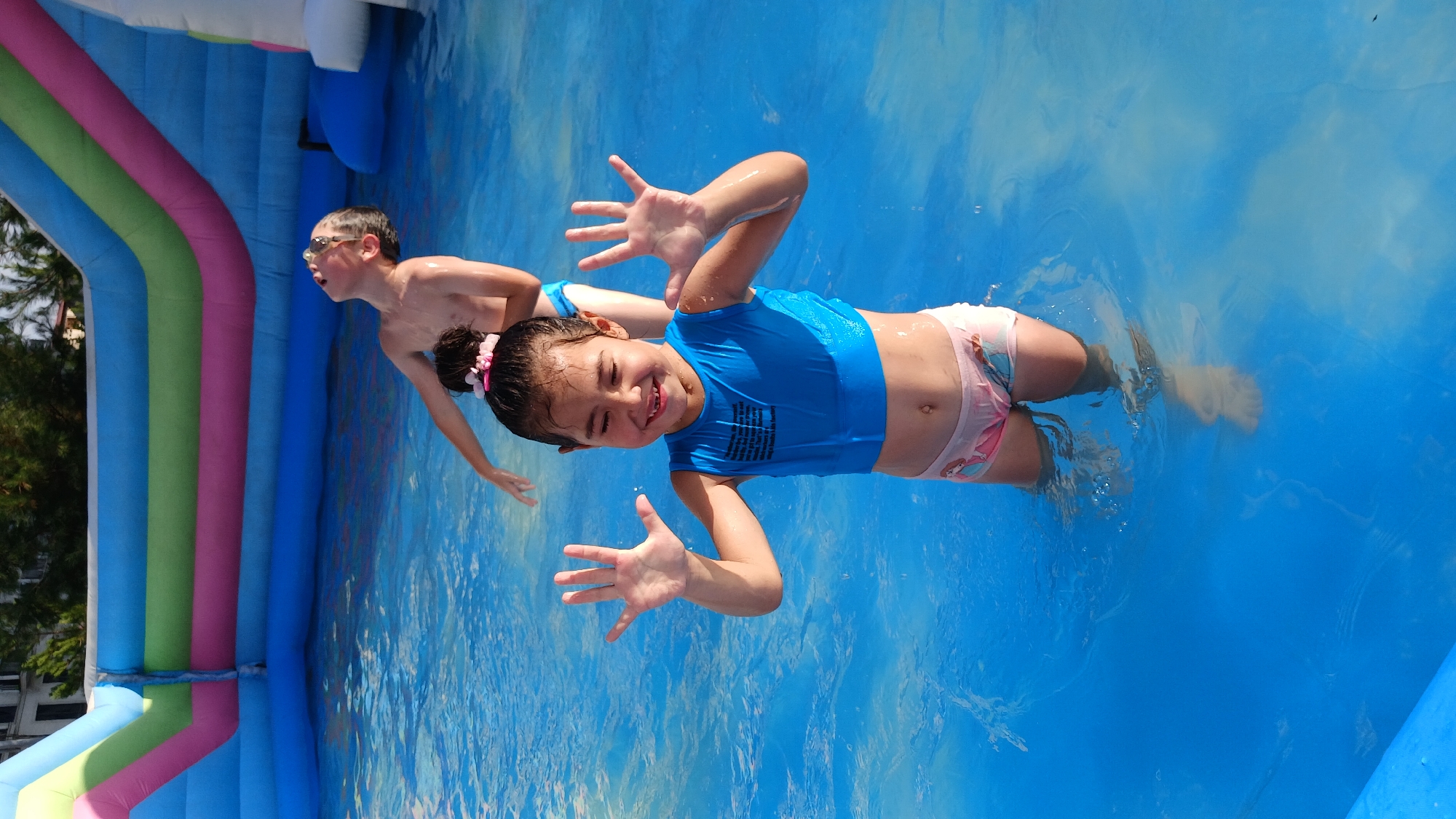 In Fergana City, Uzbekistan, two children are pictured in a brightly colored inflatable pool. A young girl in the foreground, wearing a blue swim vest and pink patterned shorts, smiles widely with her hands raised. Behind her, a boy with glasses looks upwards, his body tilted back, and he appears to be laughing. The pool is filled with clear blue water, and a large, multi-colored inflatable slide is visible in the background. The scene suggests a day of fun and recreation.